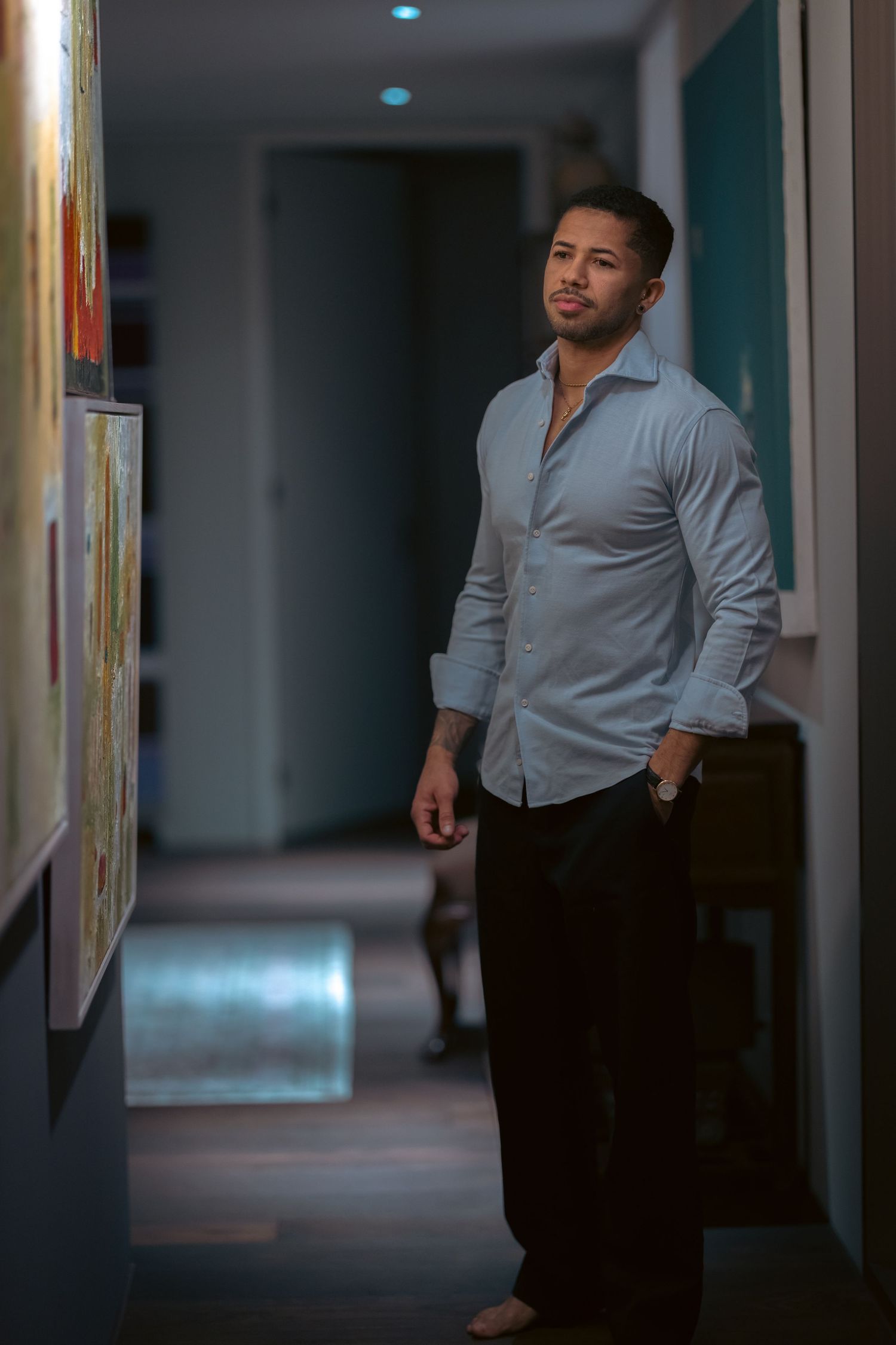 A stylish man in a light blue shirt and black pants stands in a dimly lit hallway with artwork on the walls.
