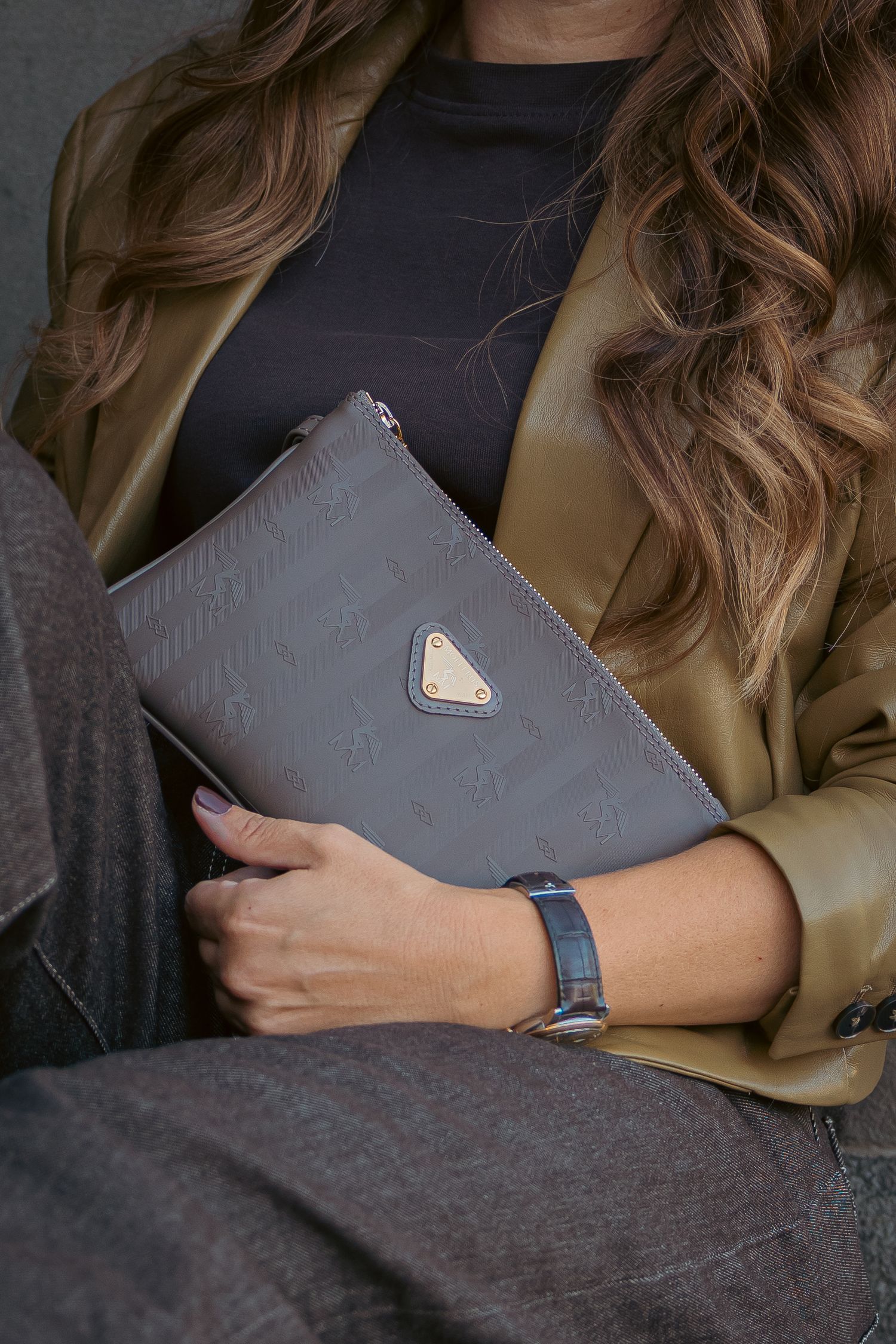 Woman holding a gray Prada monogram clutch bag, wearing a olive blazer, black top, and a dark watch.