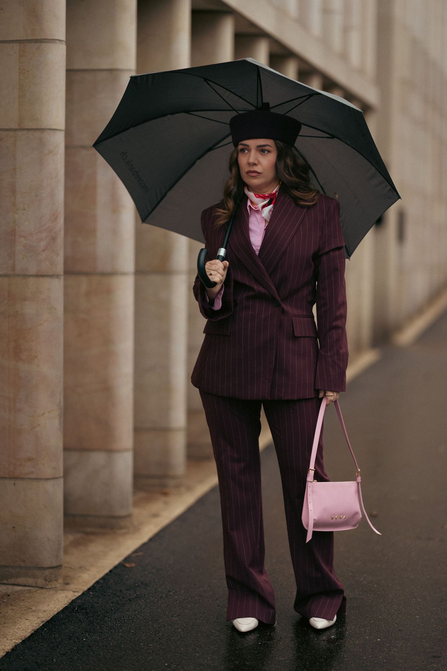 Stylish figure in burgundy pinstripe suit holding black umbrella and pink handbag stands between stone columns.