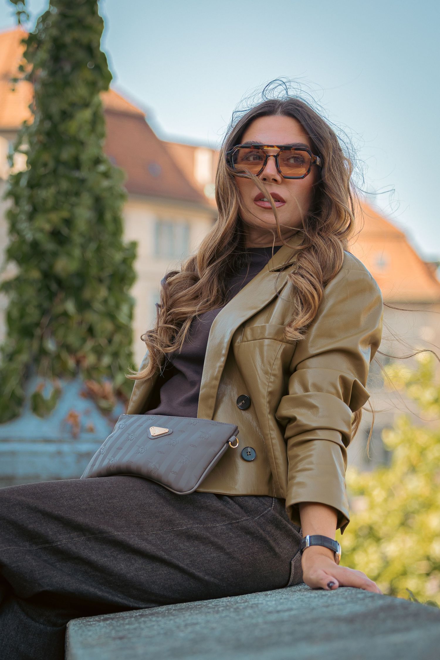 Stylish woman in beige leather jacket and sunglasses sitting outdoors with a gray clutch bag.