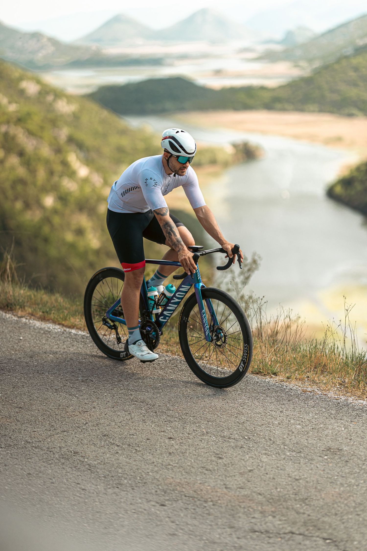 Professional cyclist riding teal Canyon road bike descending mountain road with stunning lake and valley backdrop.