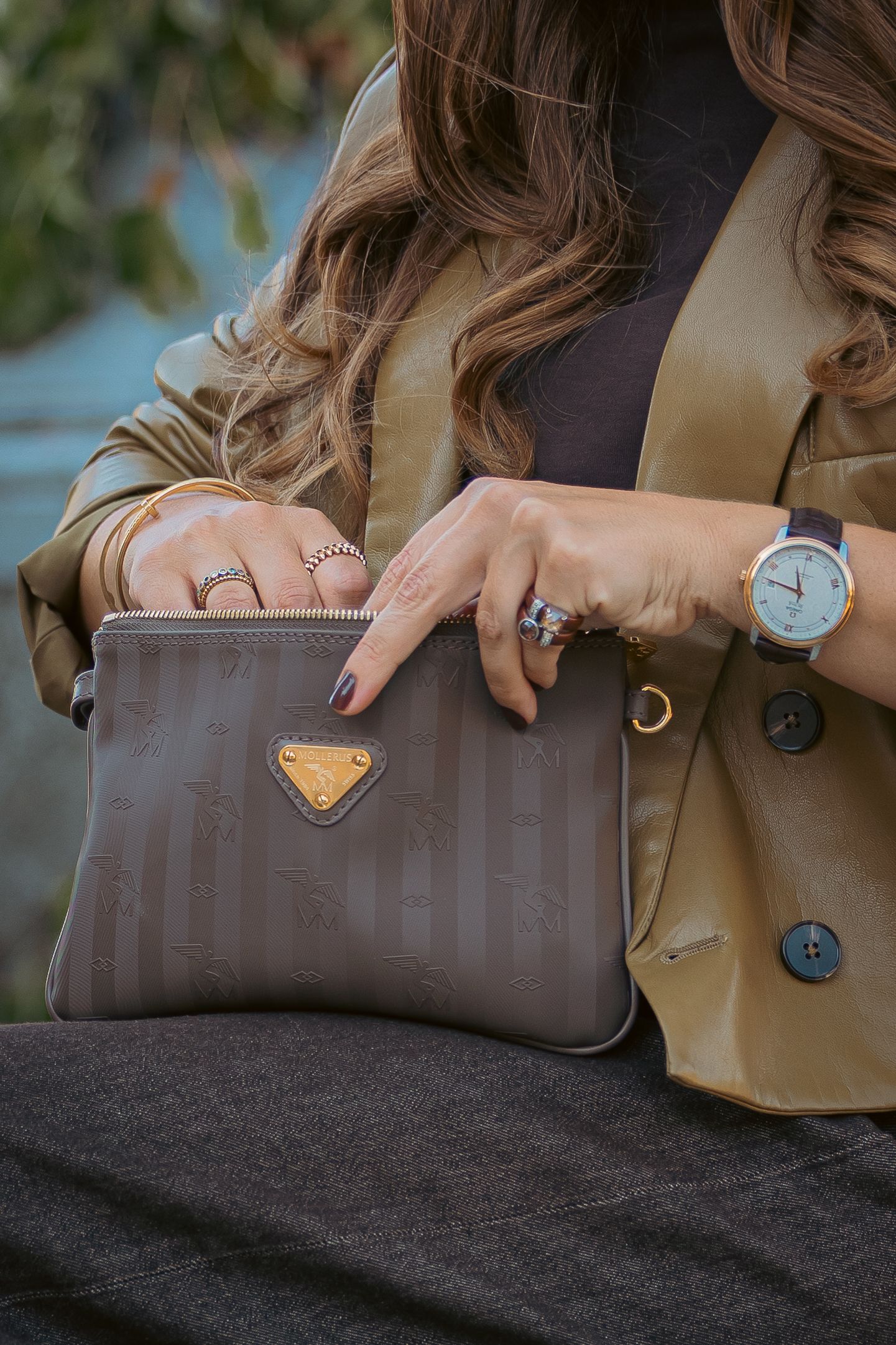 Woman holding a taupe Prada clutch bag, wearing gold rings, a brown watch, and a tan blazer.