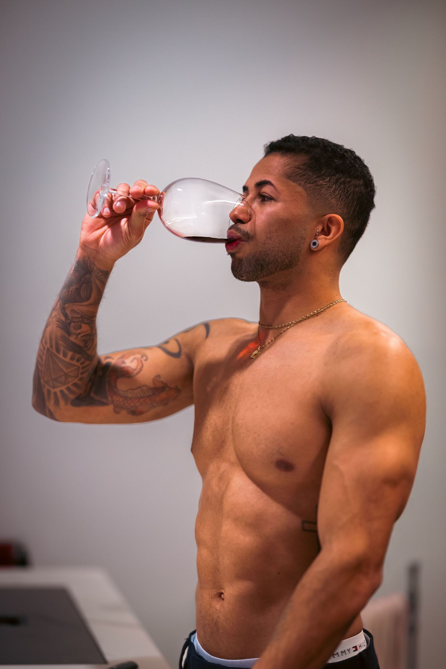 Shirtless muscular man with tattoos and gold chain drinking red wine from a large glass indoors.