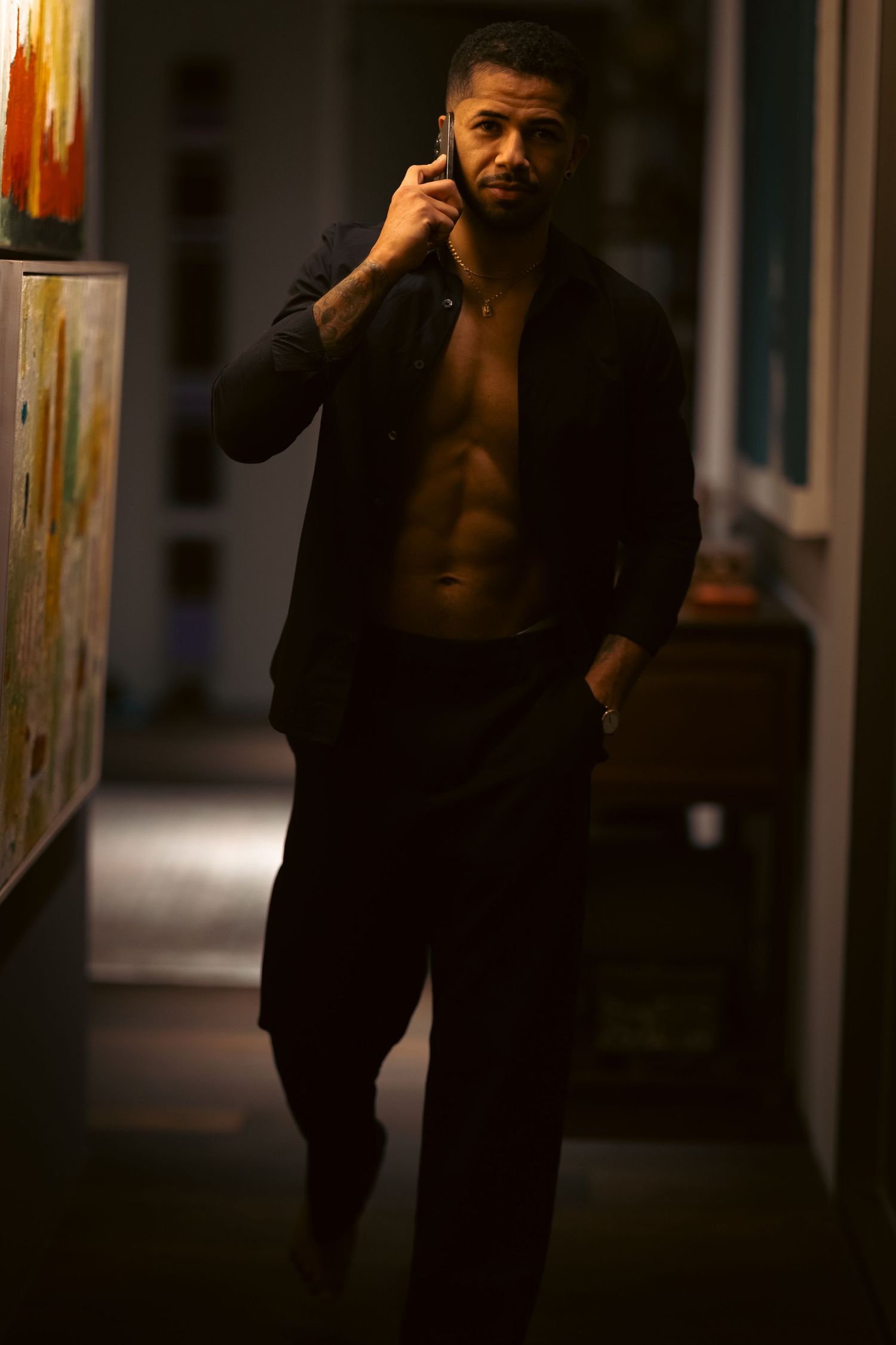 Muscular man with open black shirt talking on phone in dimly lit hallway with colorful artwork.