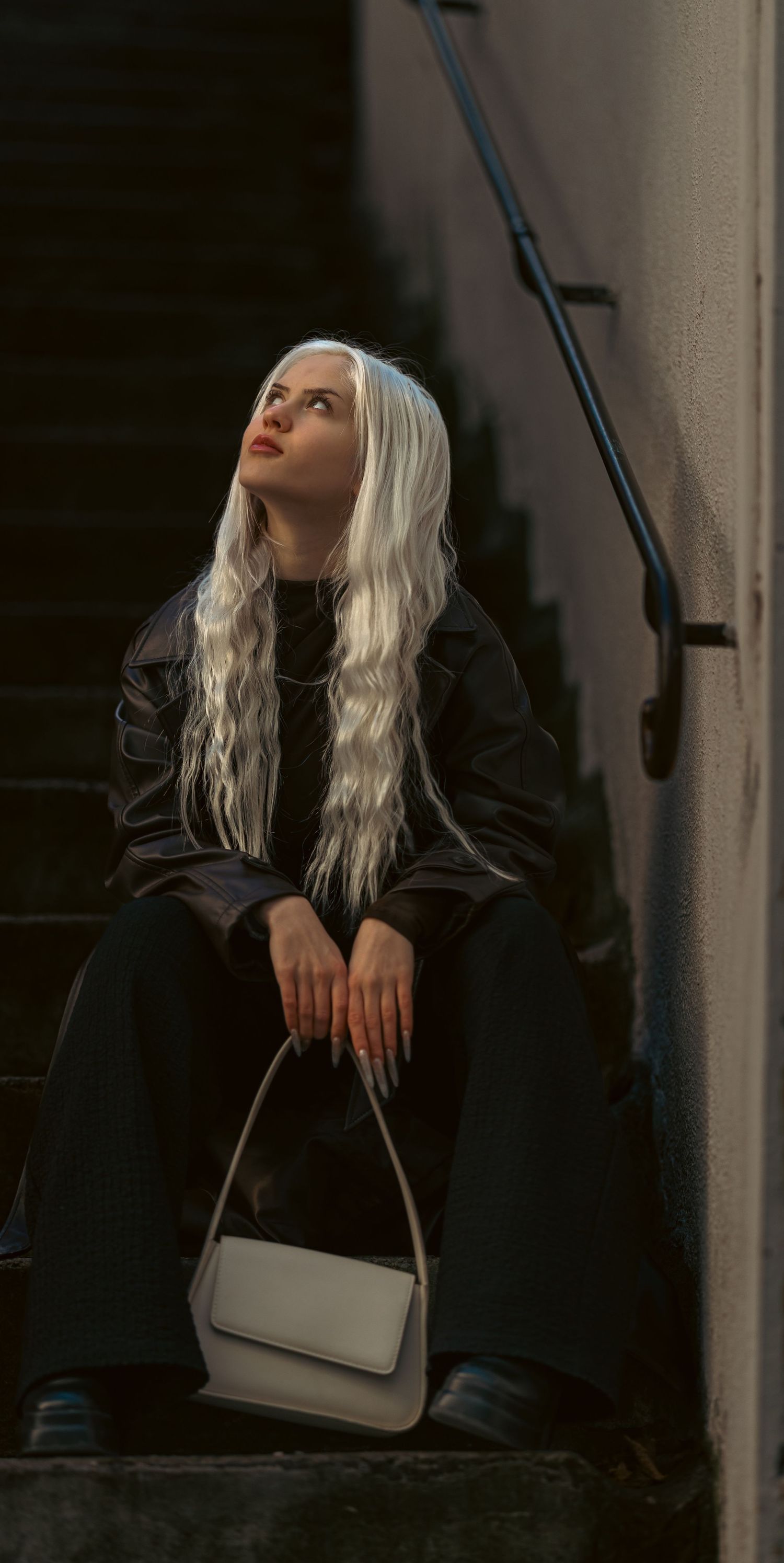 Young woman with long white hair sits on dark stairs wearing black leather jacket, holding a white handbag, looking up.