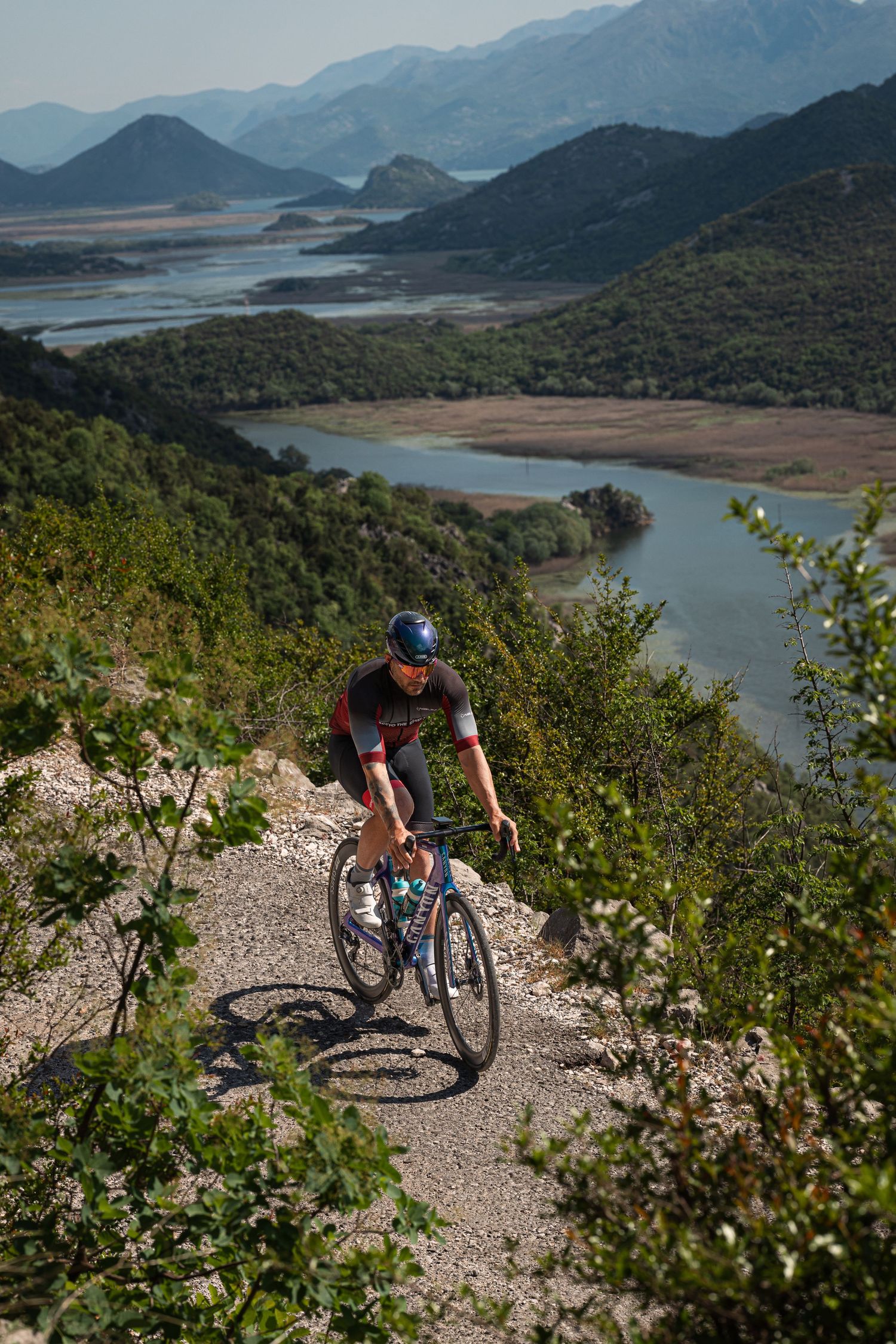 Cyclist riding uphill on a rocky mountain trail with scenic river valley and green hills in the background.