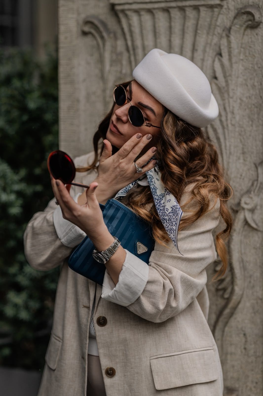 Stylish woman in white beret and sunglasses checks makeup in compact mirror outdoors.