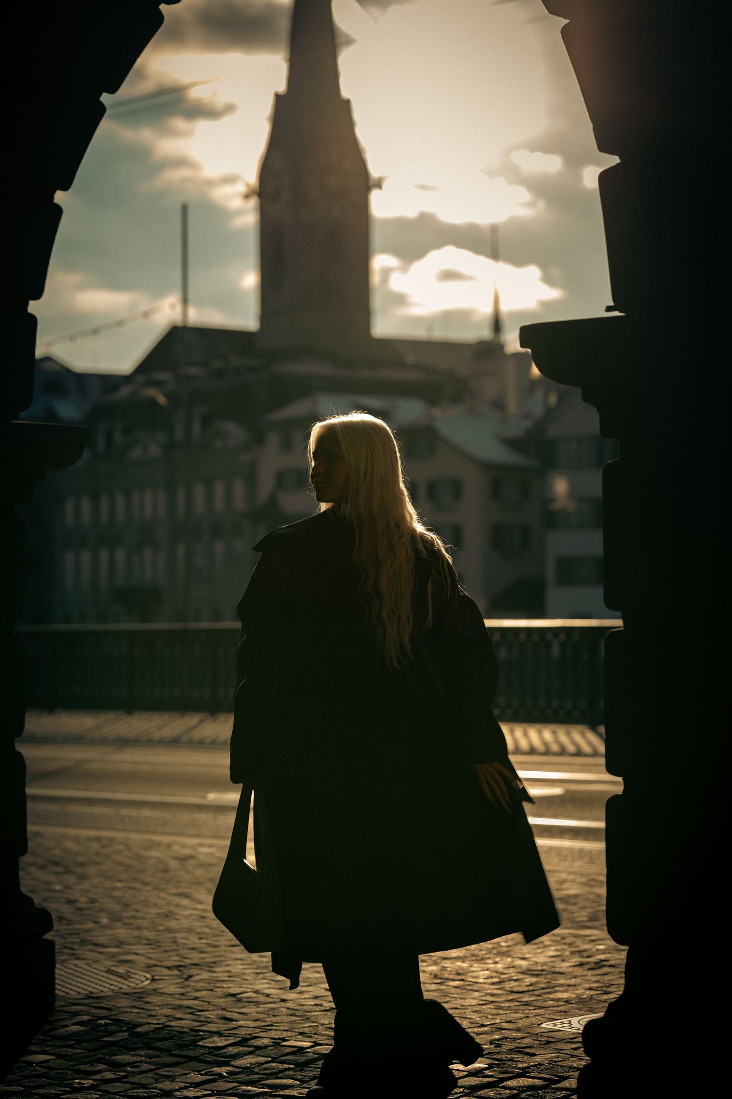 Silhouetted figure walks across cobblestone street at sunset with city buildings in background creating moody atmosphere.
