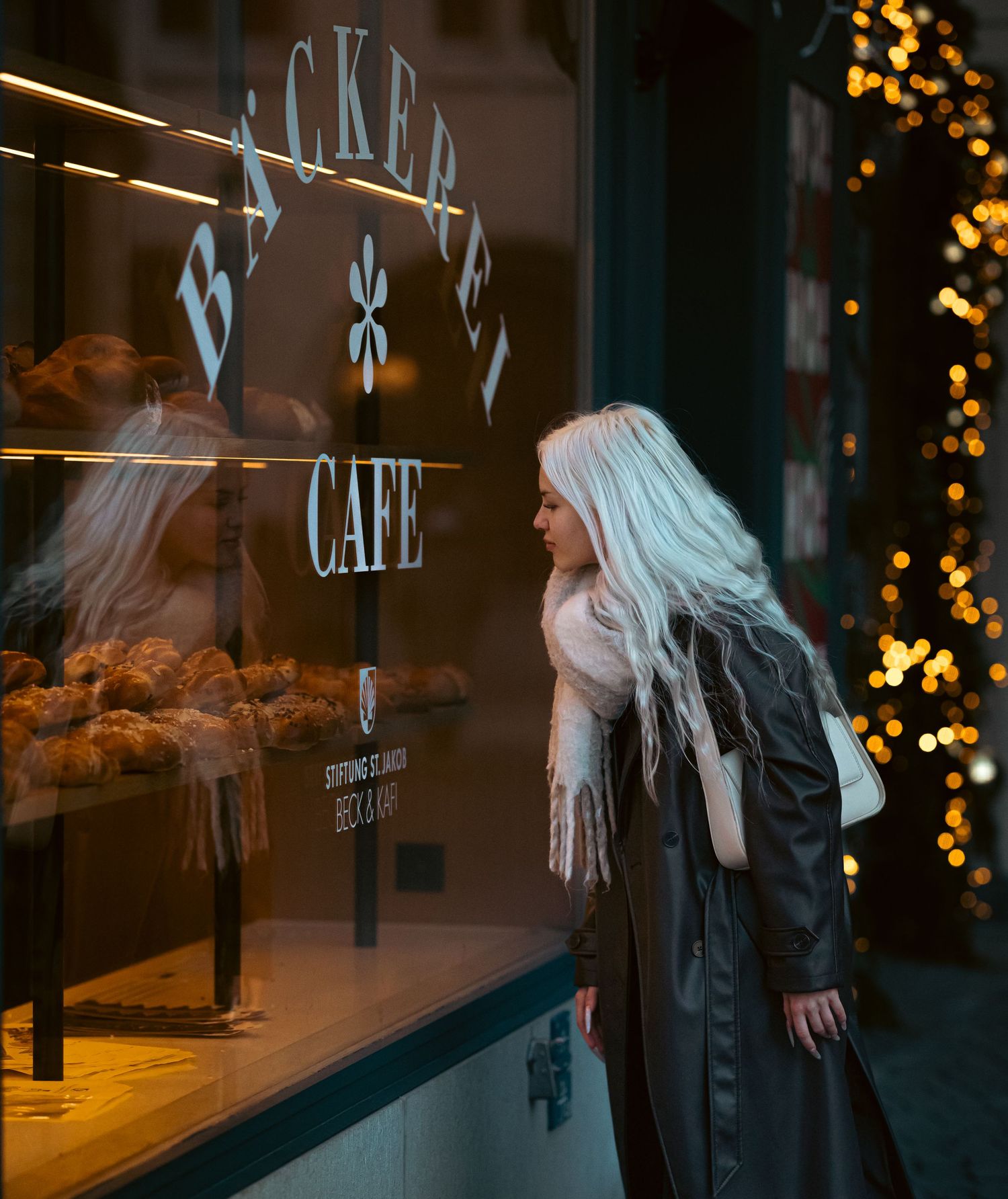 Person in black coat stands near illuminated cafe window with blurred bokeh lights at night.