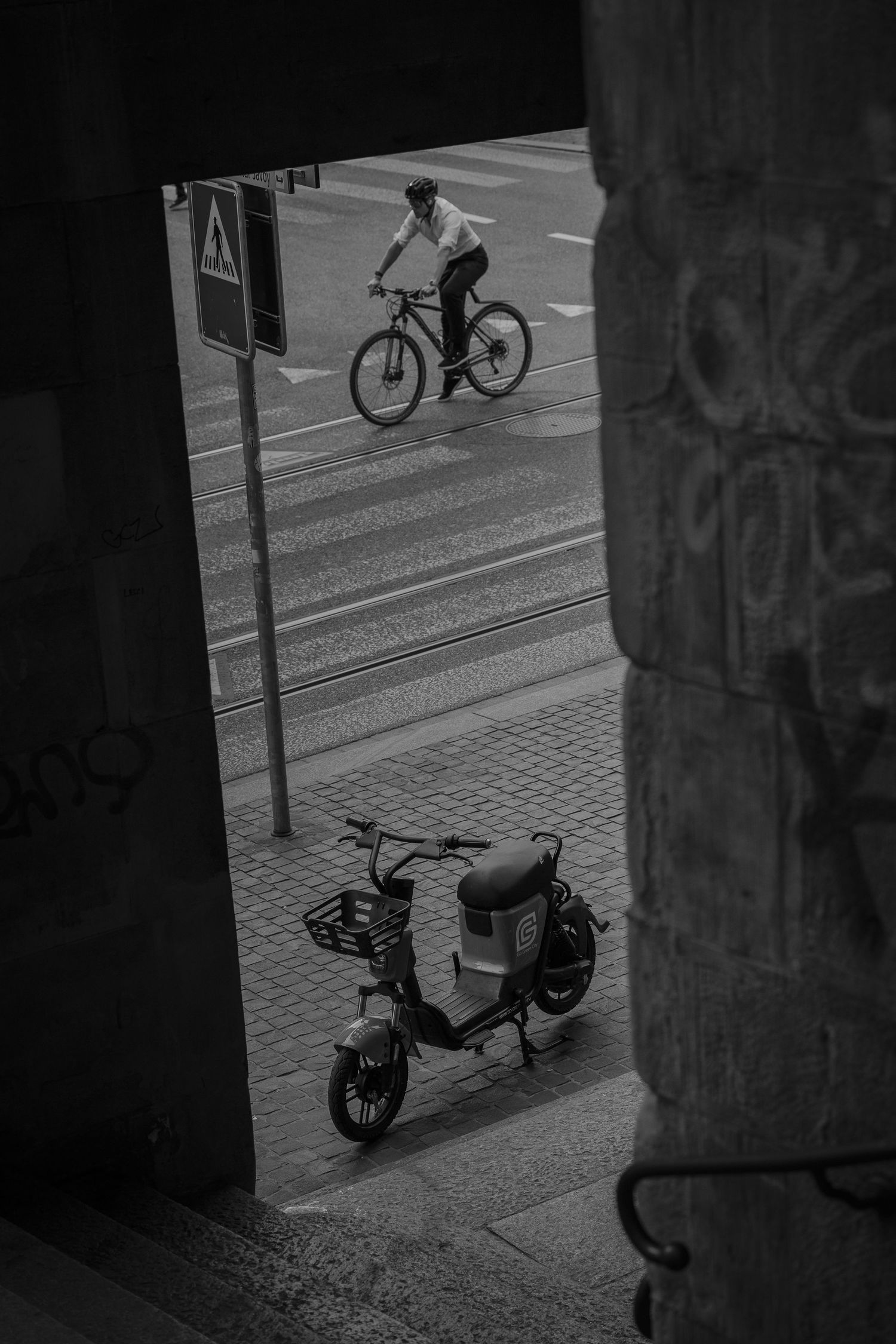 Black and white urban street photo showing a cyclist riding past tram tracks while an electric scooter sits in the foreground.