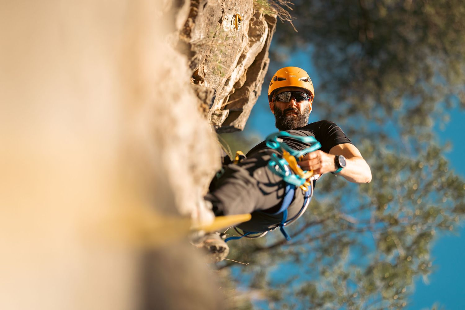 A rock climber in an orange helmet and harness scales a steep cliff face against a bright blue sky.