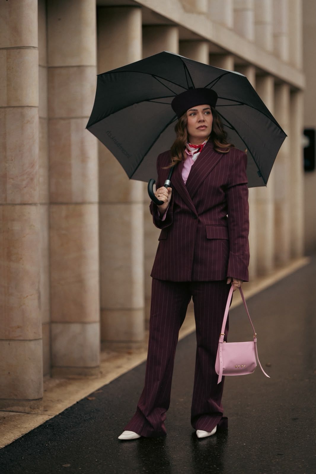 Woman in burgundy pinstripe suit holding black umbrella and pink bag, wearing beret outdoors.
