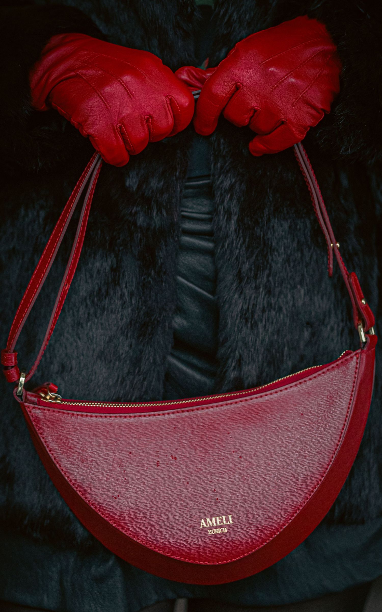 Red leather AMELI Zurich crescent handbag held by hands in red gloves against a dark fur coat.