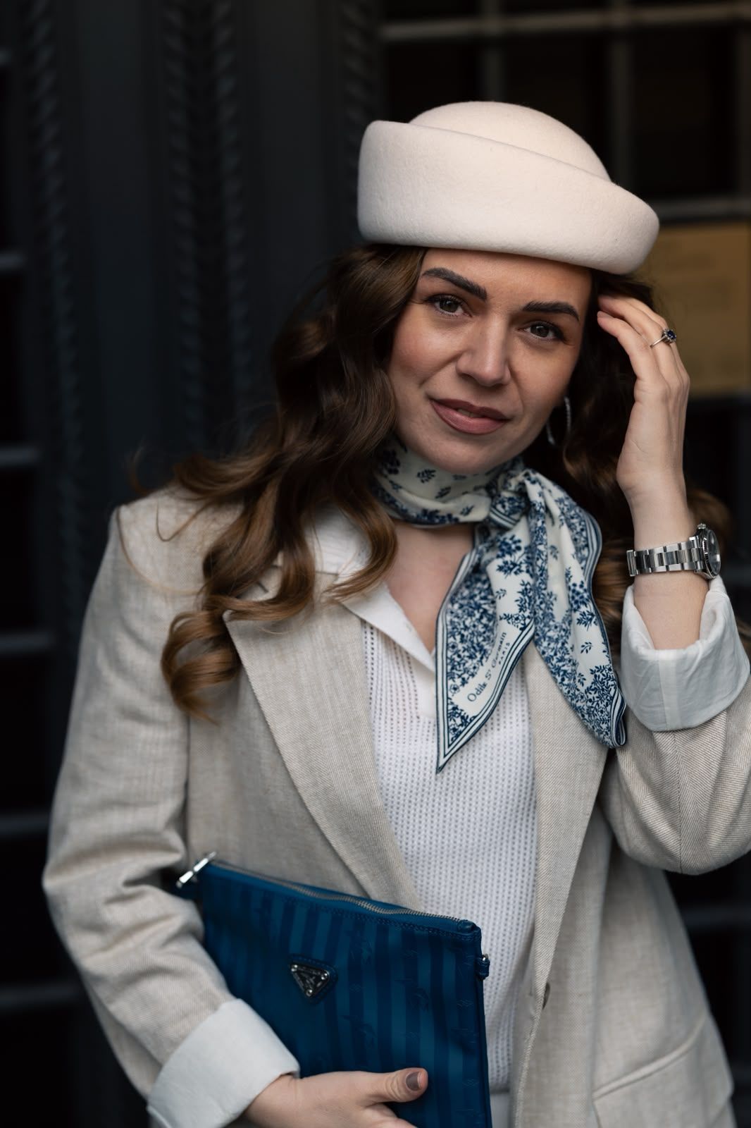 Stylish woman in beige beret, scarf, and blazer holding a blue clutch bag outdoors.