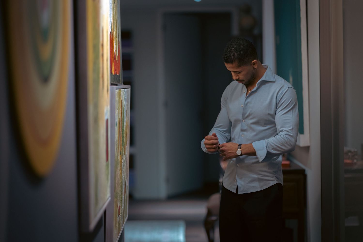 Man in light blue shirt buttoning cuffs in a hallway with colorful artwork on the walls.