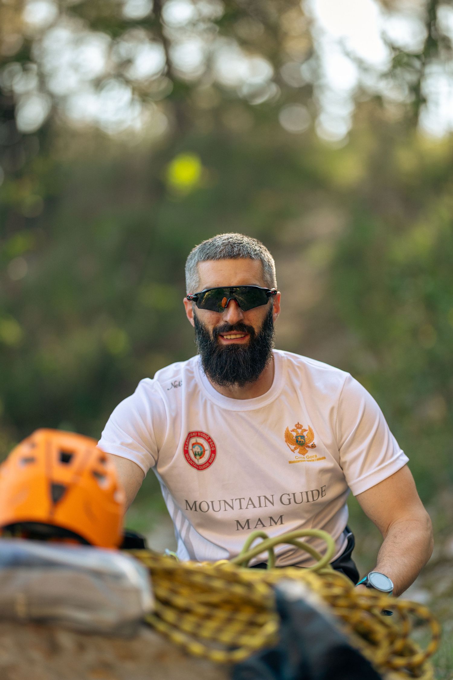 Bearded mountain guide in white MAM t-shirt and sunglasses prepares climbing ropes outdoors.