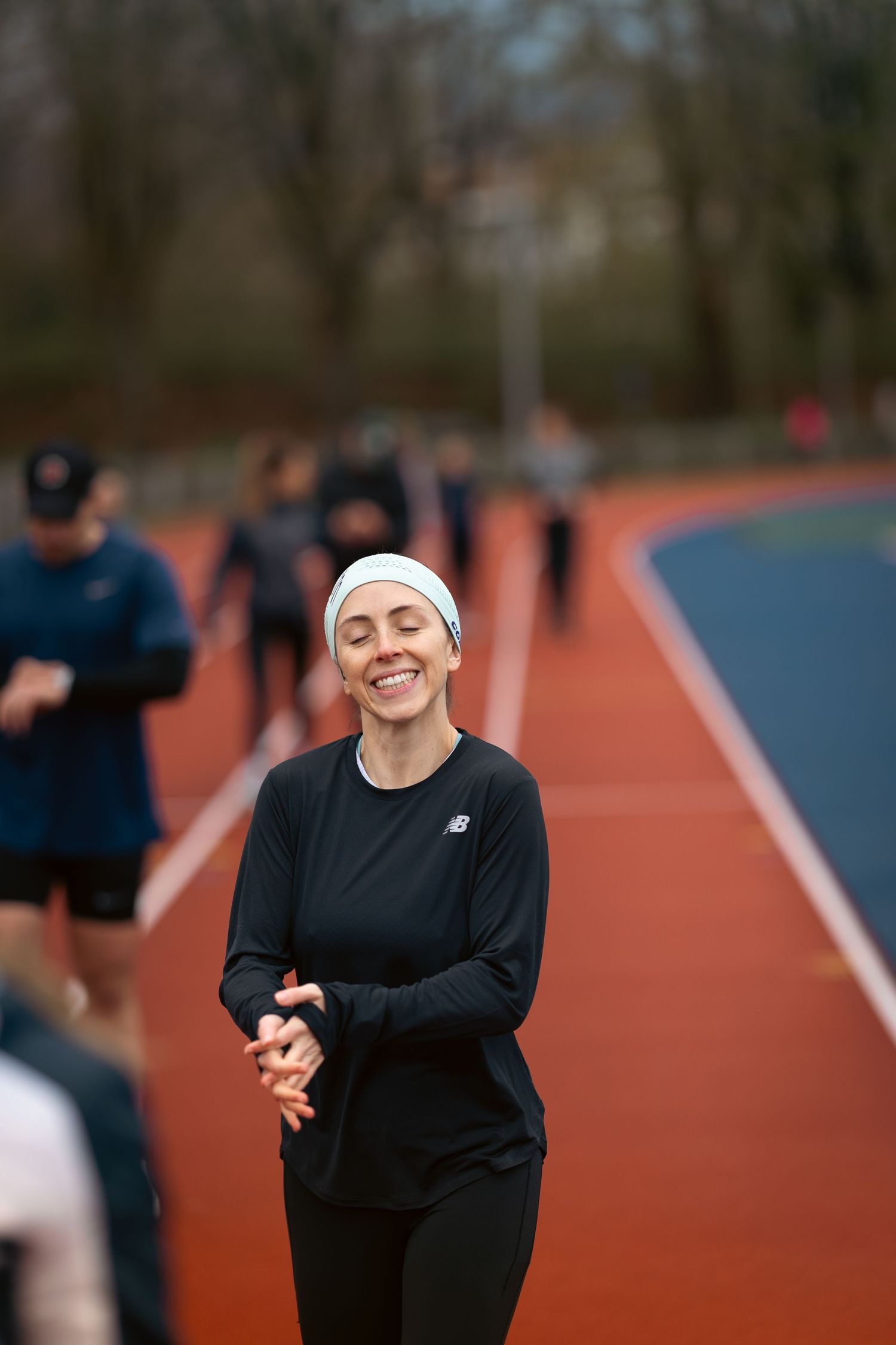 Smiling woman in black New Balance running gear and white headband jogging on a red athletic track.