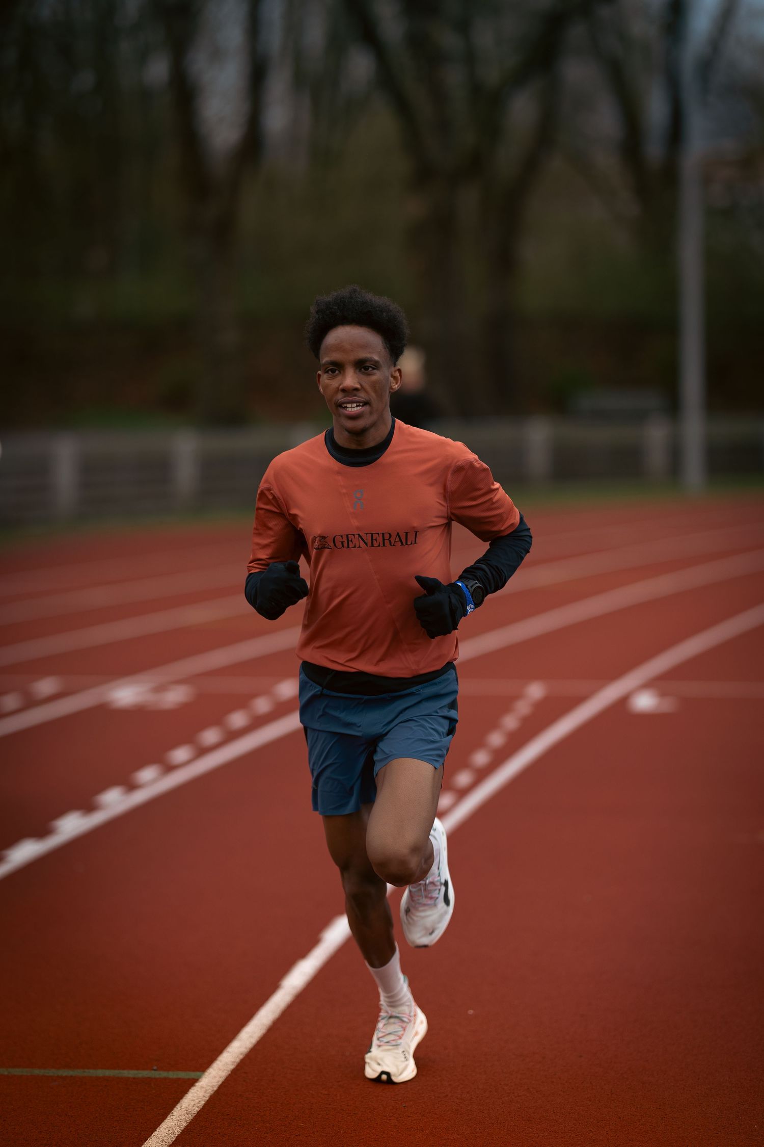 Young male athlete running on a red track wearing an orange long-sleeve shirt, blue shorts, and white sneakers.