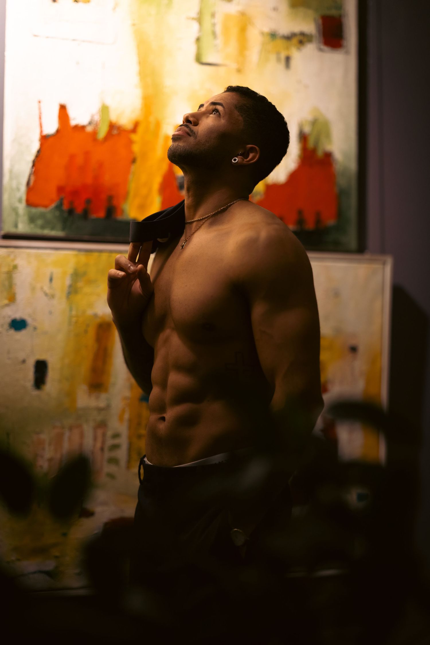 A shirtless athletic man with defined abs posing in front of colorful abstract artwork.