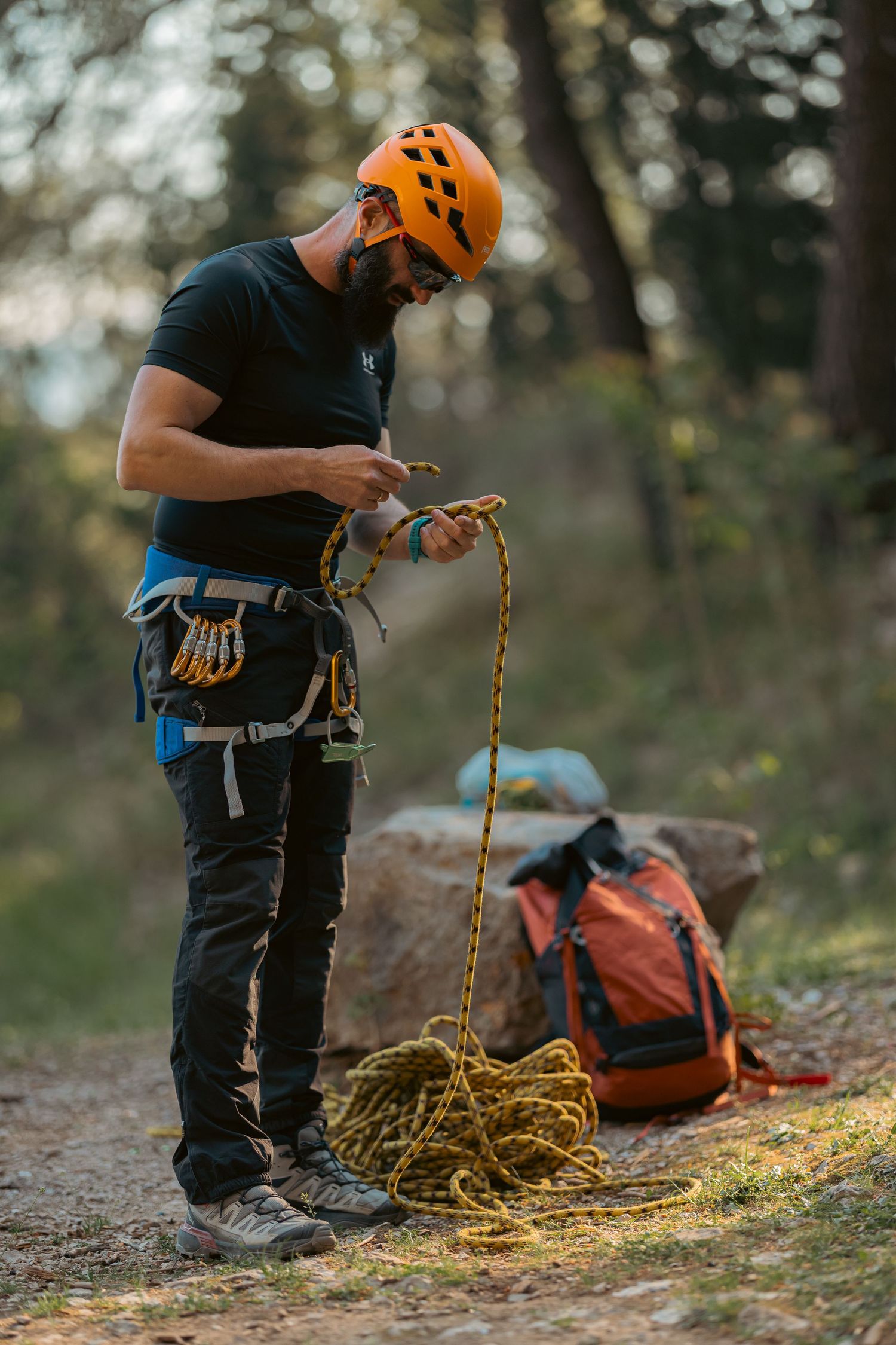 Rock climber in orange helmet preparing rope and gear in forest setting before a climb.