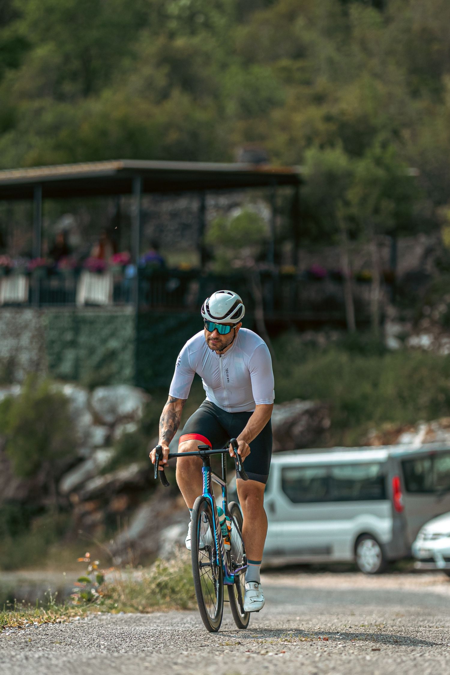 A male cyclist in white jersey and helmet rides a blue road bike uphill on a paved road with green hills behind.