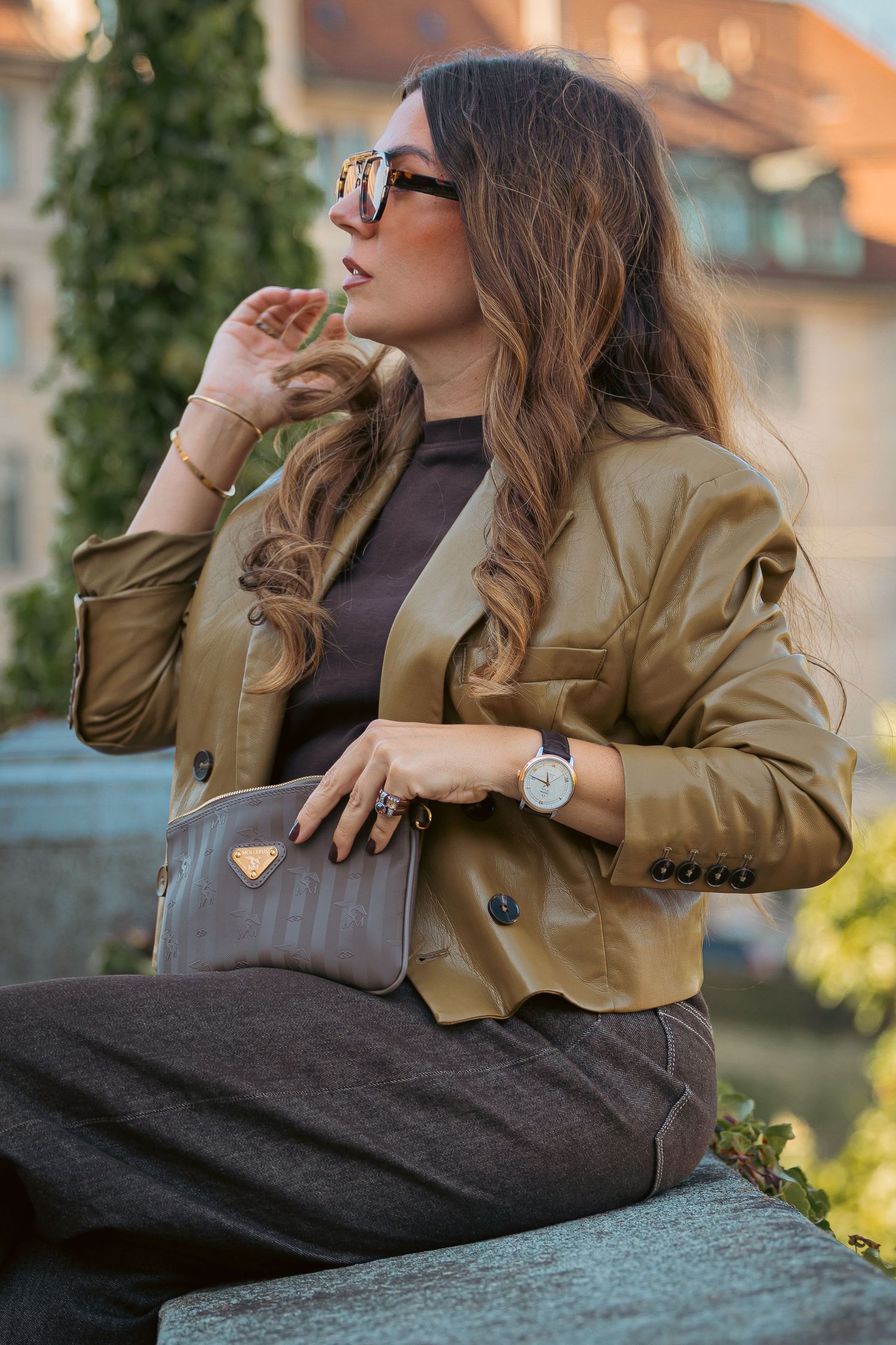 Stylish woman in tan leather jacket, sunglasses, and gray trousers holding a brown bag outdoors.