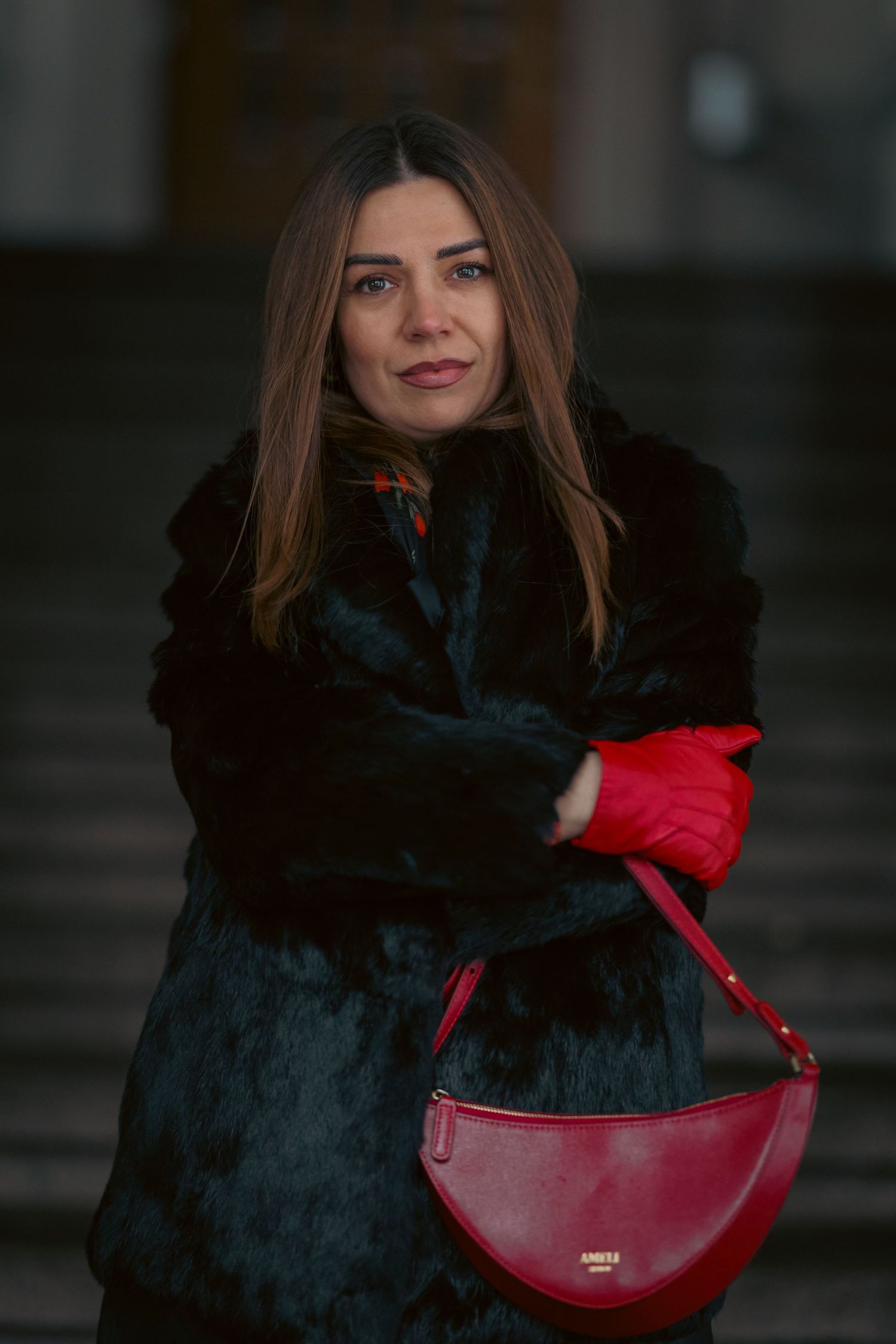 A person in a black fur coat poses with a red leather handbag against a dark background.