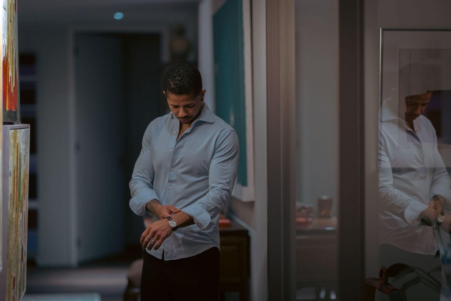 Man in light blue shirt checking his watch in a modern hallway with mirror reflection.