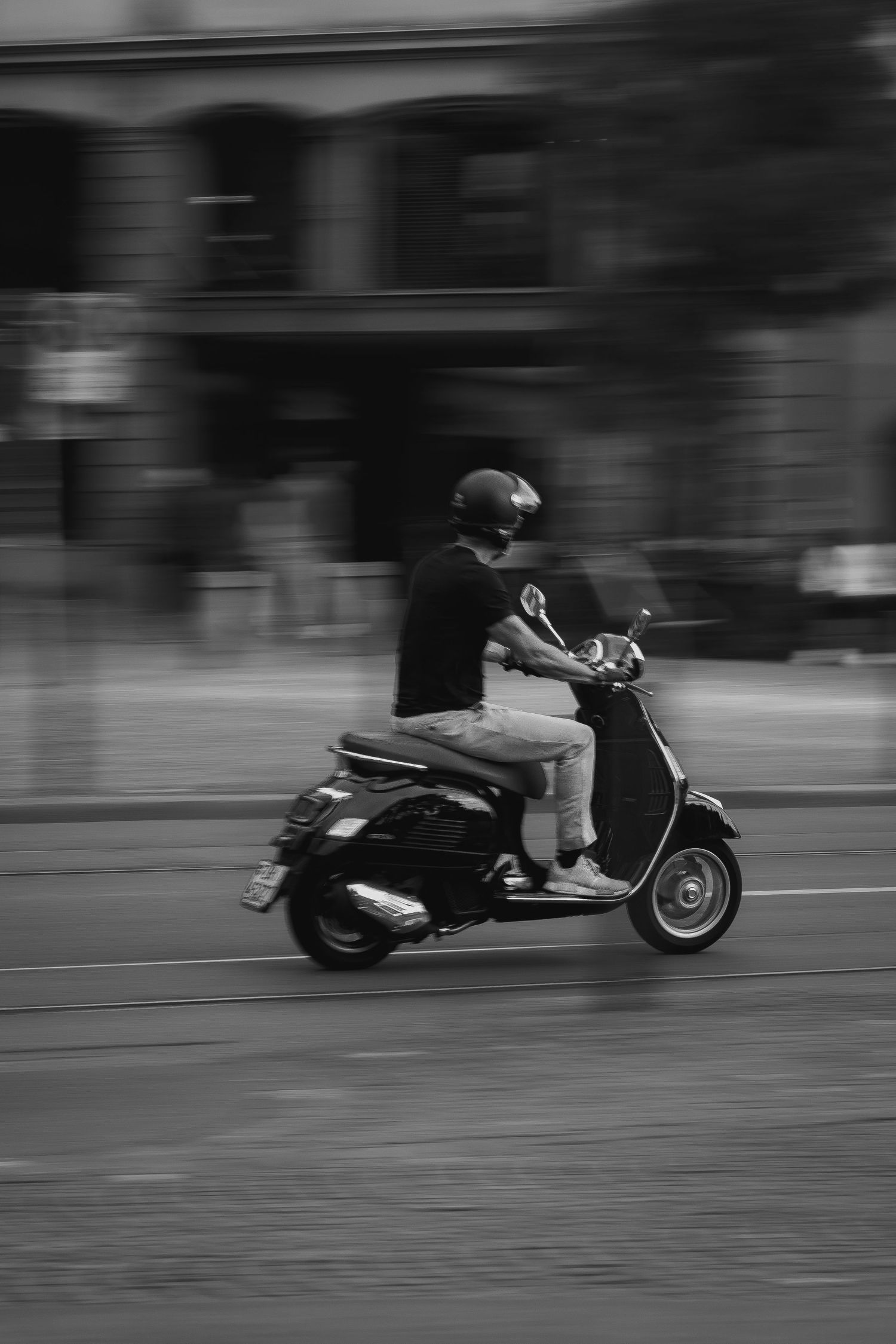 Black and white panning shot of a helmeted rider on a classic Vespa scooter speeding through city streets.
