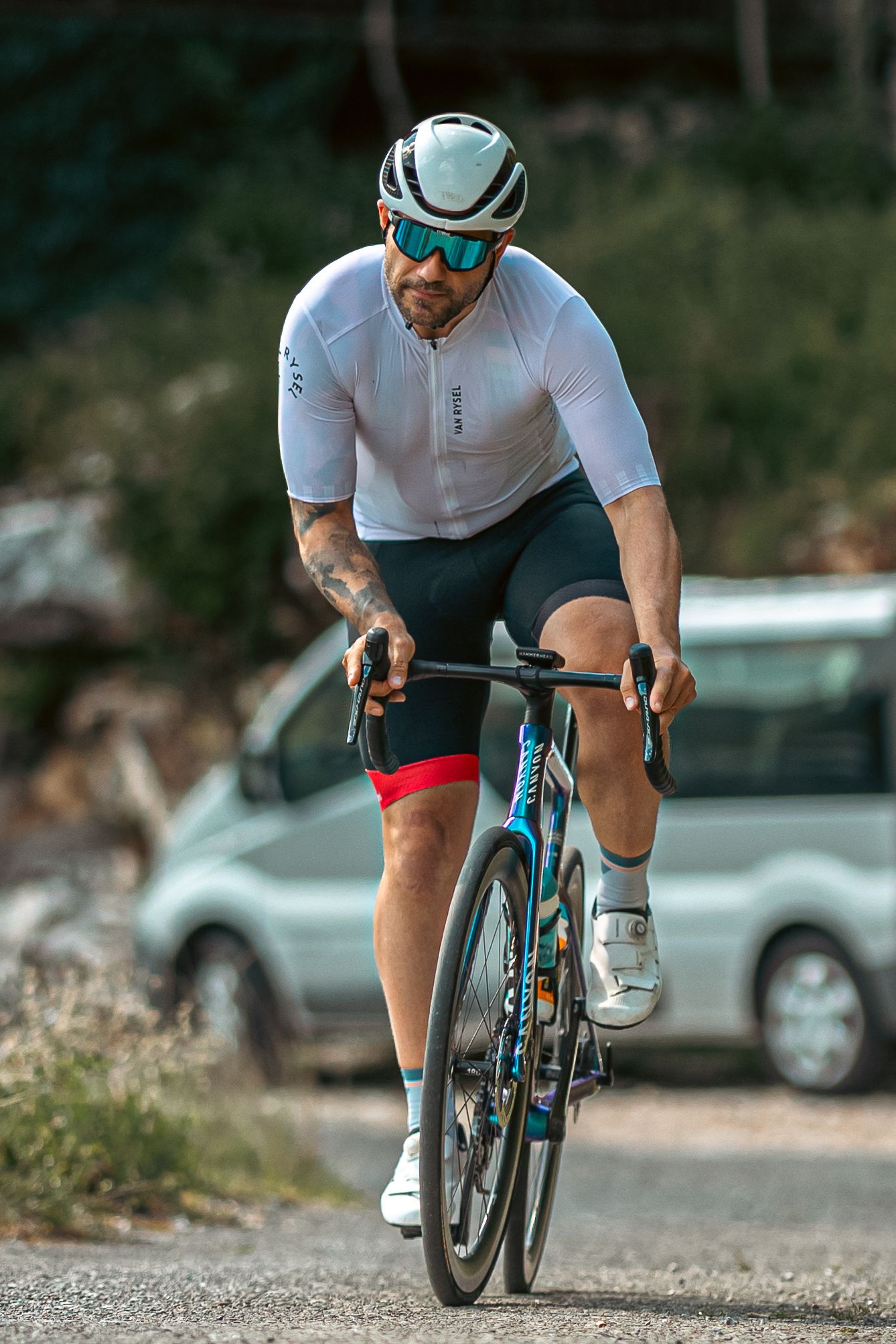 Male cyclist in white jersey and helmet riding a road bike on a paved path with cars in the background.