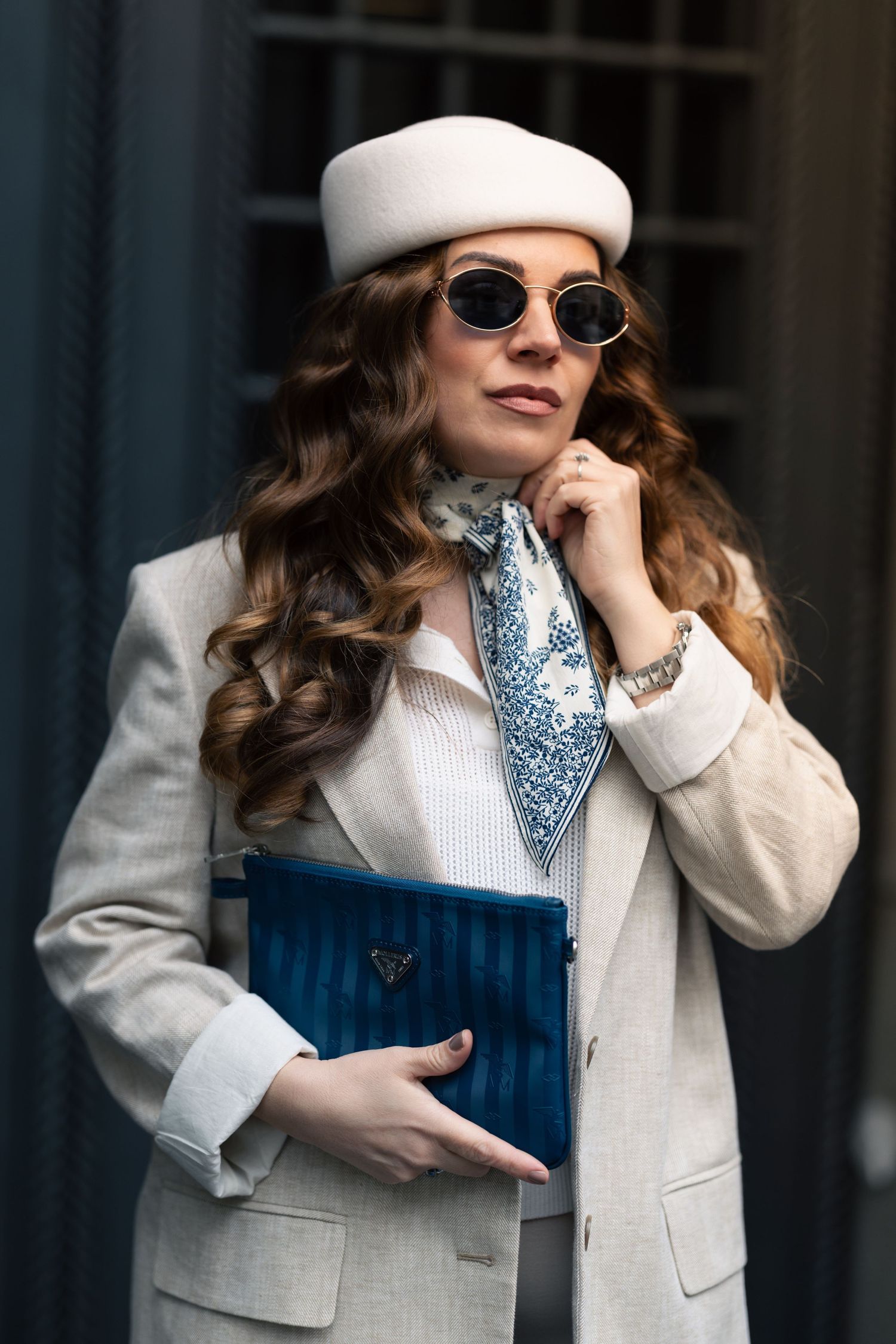 Fashionable woman in white coat and beret with sunglasses holds blue clutch while posing against dark building exterior.