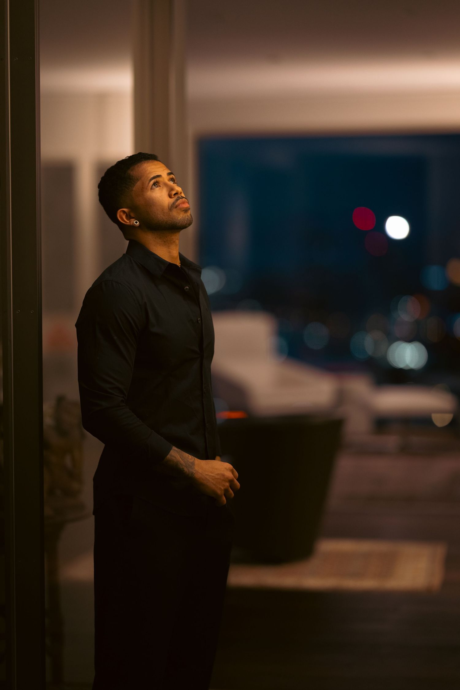 Athletic man in black outfit standing by window at night with city lights bokeh background.