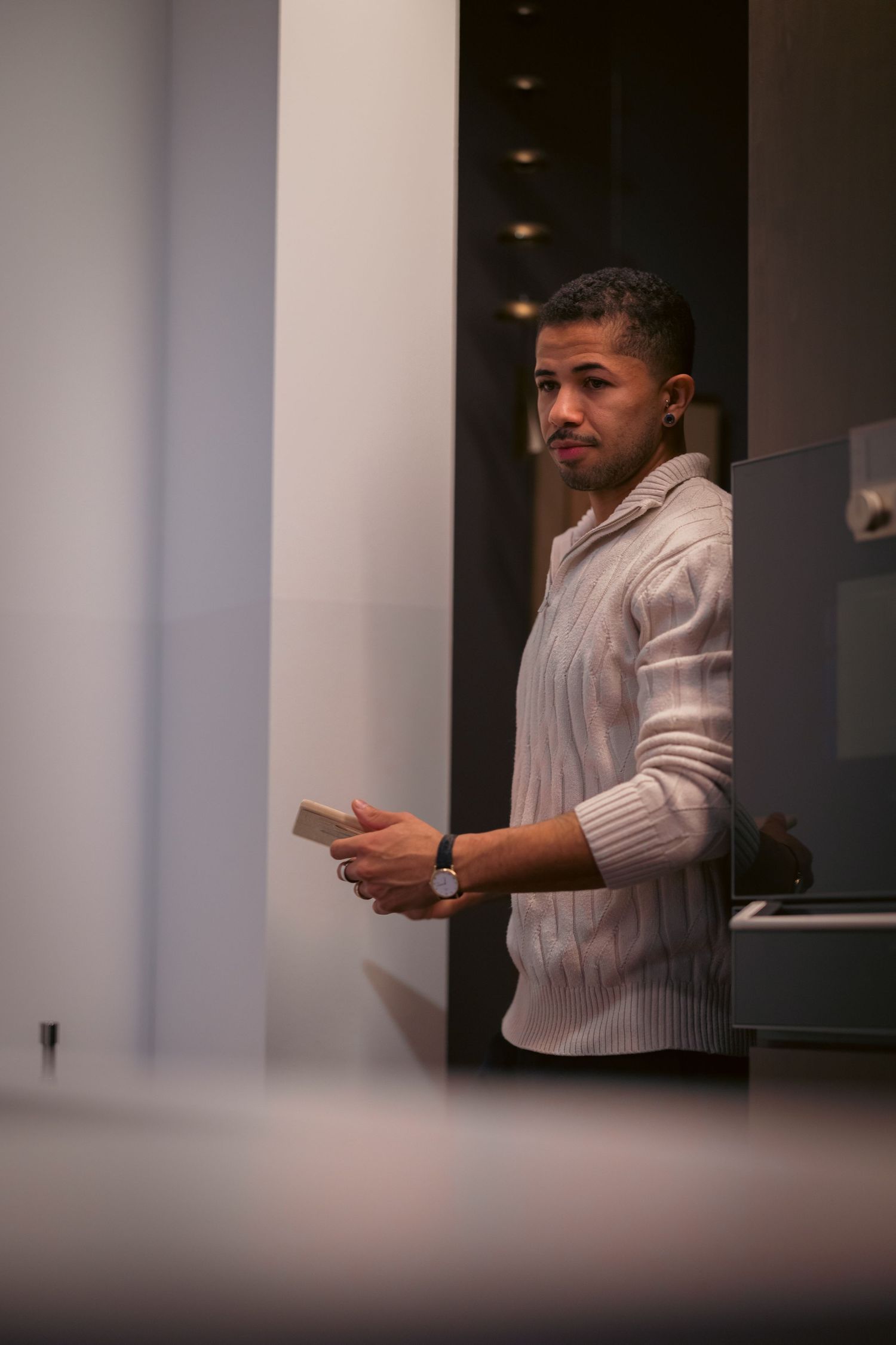 A young man in a white knit sweater holding a card while standing in a modern interior space.