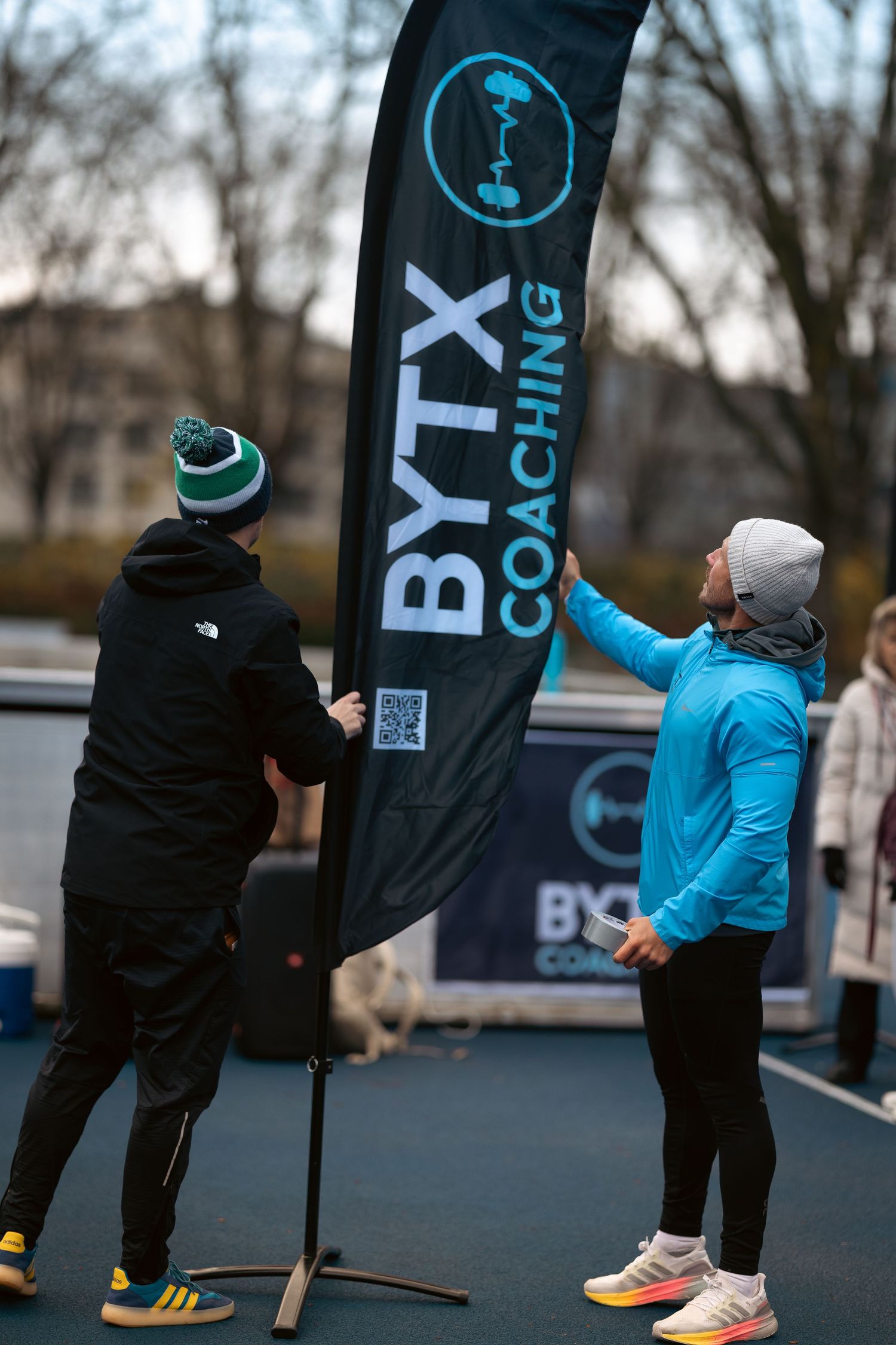 Two people setting up a black BTX Coaching banner flag at an outdoor event.