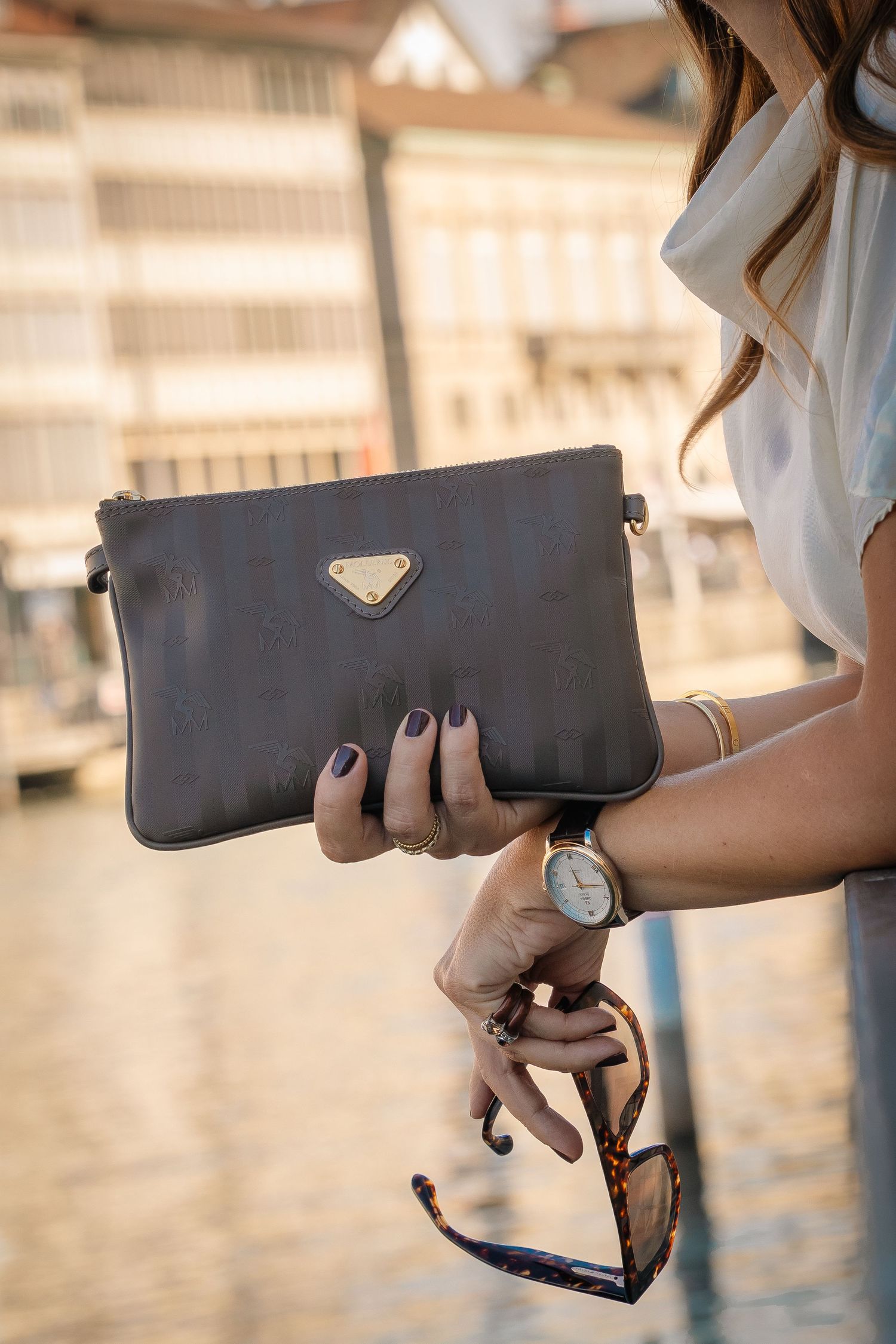 Woman holding a dark brown Prada clutch bag and sunglasses, wearing a watch and bracelet outdoors.