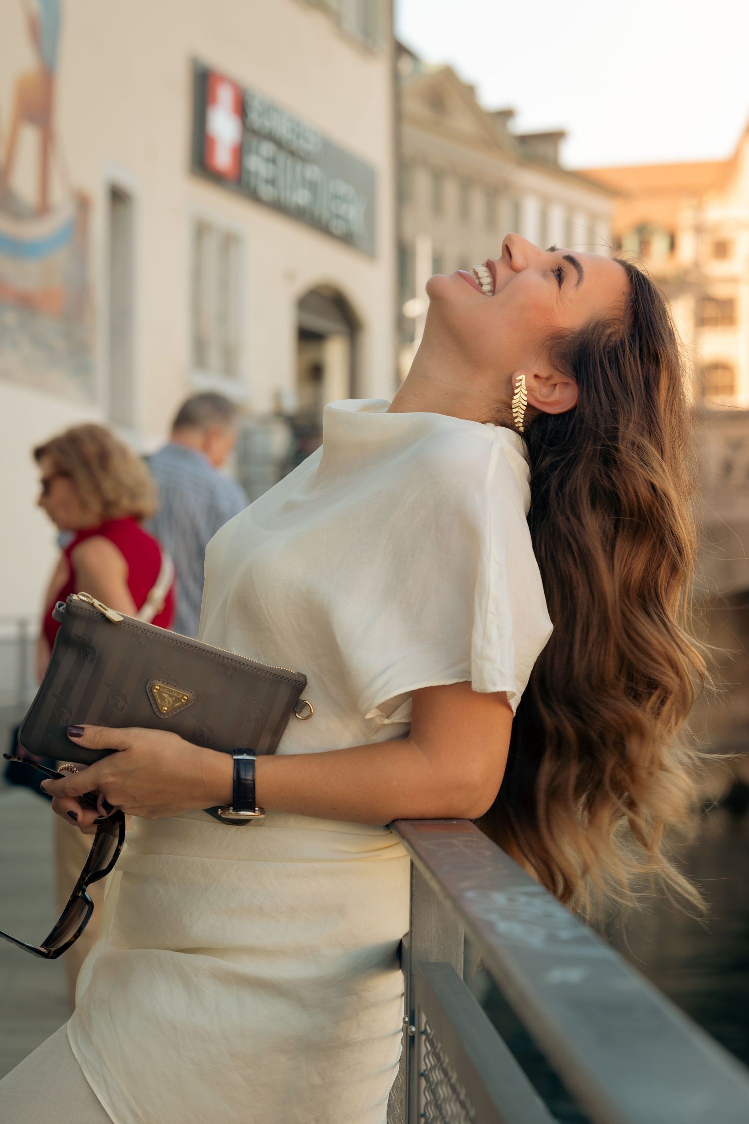 A joyful woman in a white outfit holds a Prada clutch, laughing outdoors in a European city at golden hour.