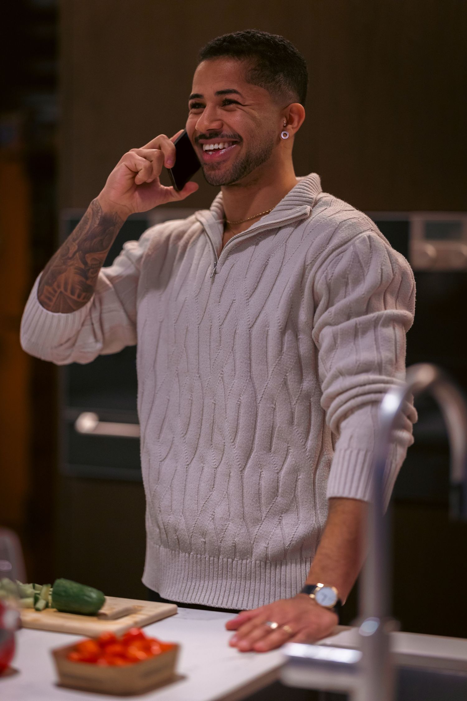 Smiling man talking on phone in kitchen, wearing cream cable-knit sweater, with fresh vegetables on counter.