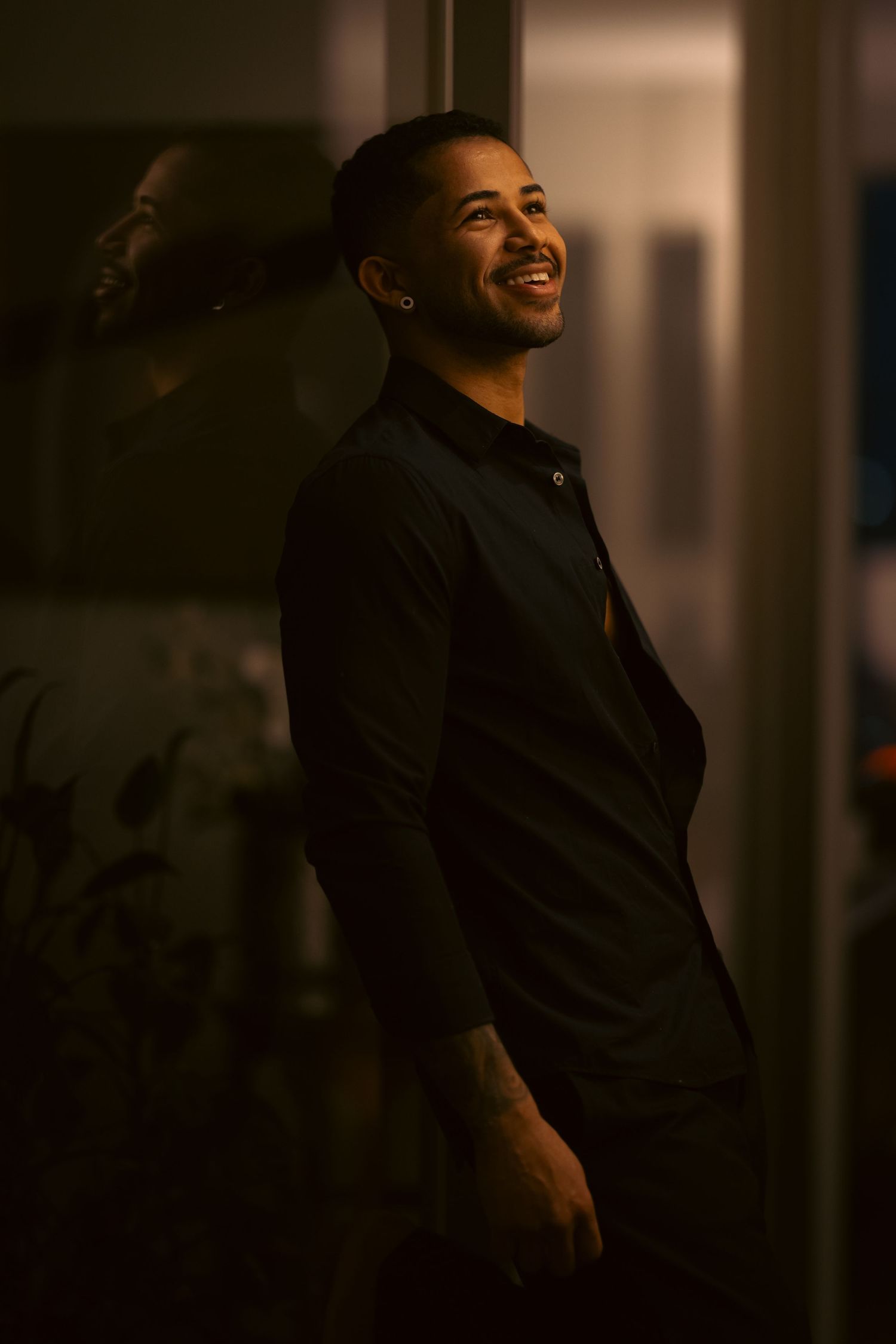Smiling young man in black shirt leaning against glass door in warm golden light.