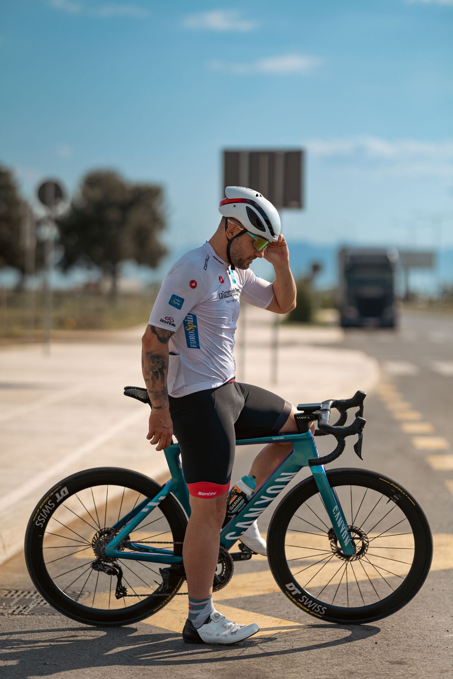 Professional cyclist in white jersey and teal Canyon bike poses on sunny road wearing white helmet and bib shorts.