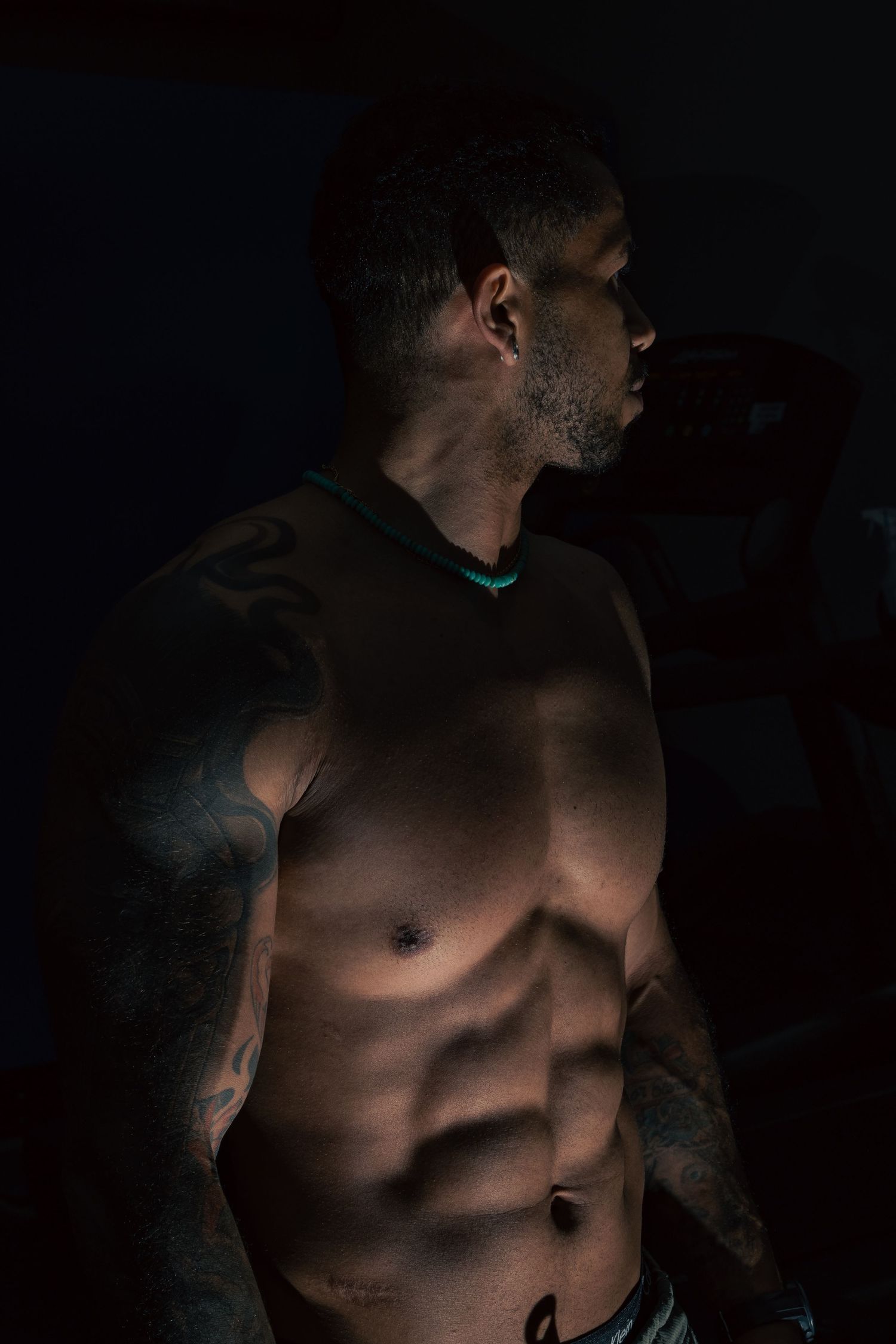 Muscular shirtless man with tattoos poses in dramatic low-key lighting, showcasing defined abs and arm tattoos.