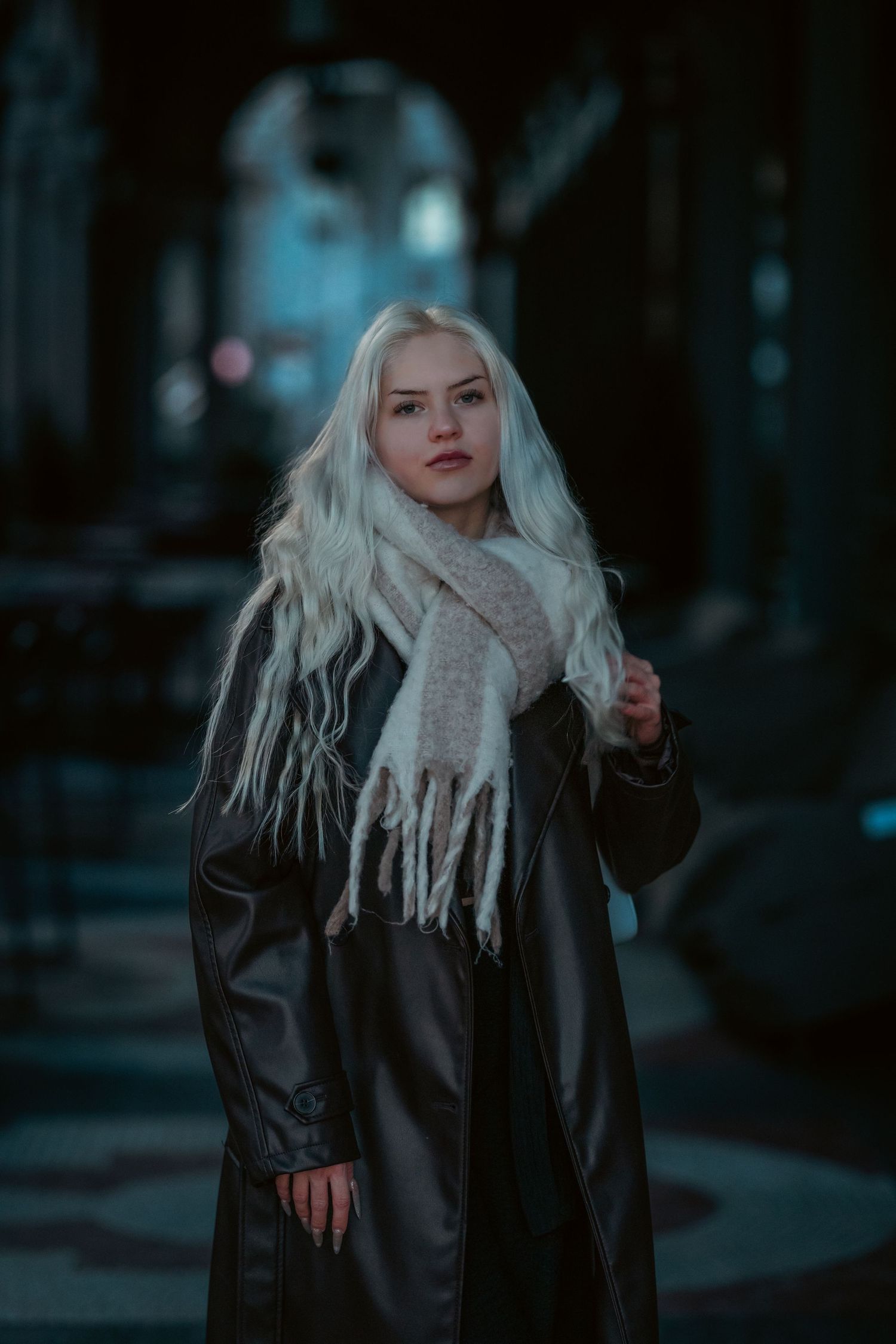 A person with long platinum blonde hair wearing a black leather coat and beige scarf stands in a moody dark setting.