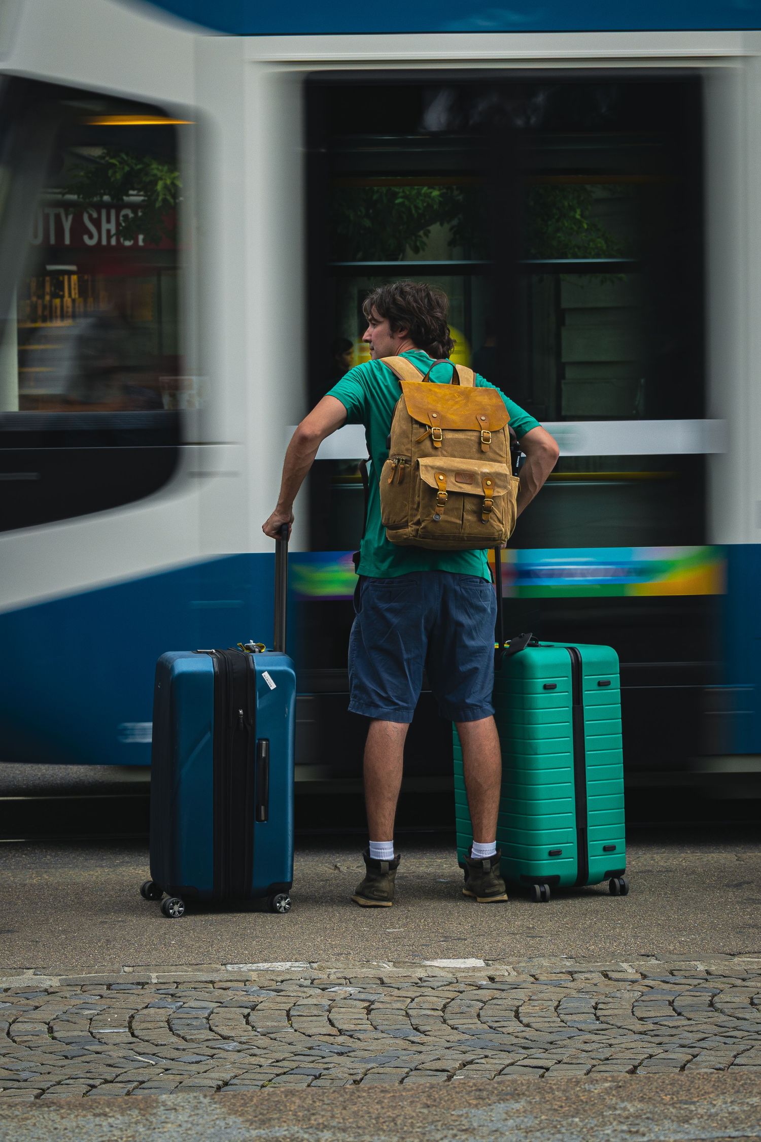 Young male traveler with backpack standing beside two suitcases at a bus stop on cobblestone street.