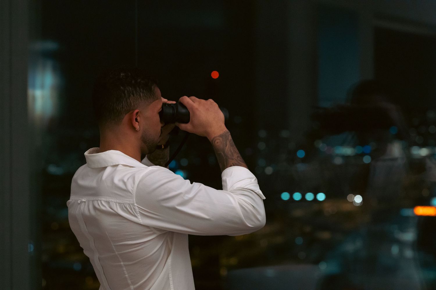 Man in white shirt using binoculars at night with city lights bokeh background.