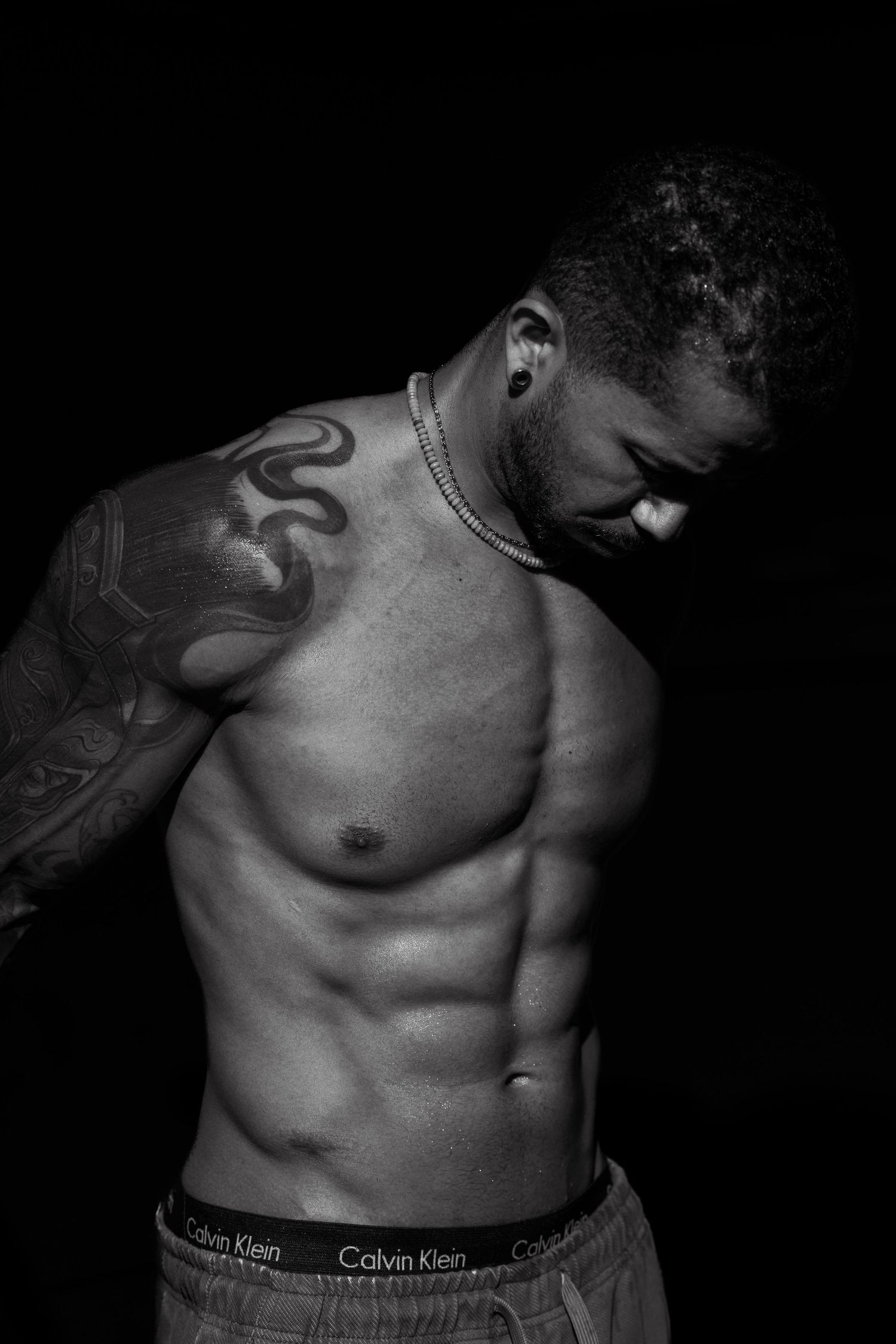 Black and white photo of a muscular shirtless man with tattoos wearing Calvin Klein underwear posing against dark background.