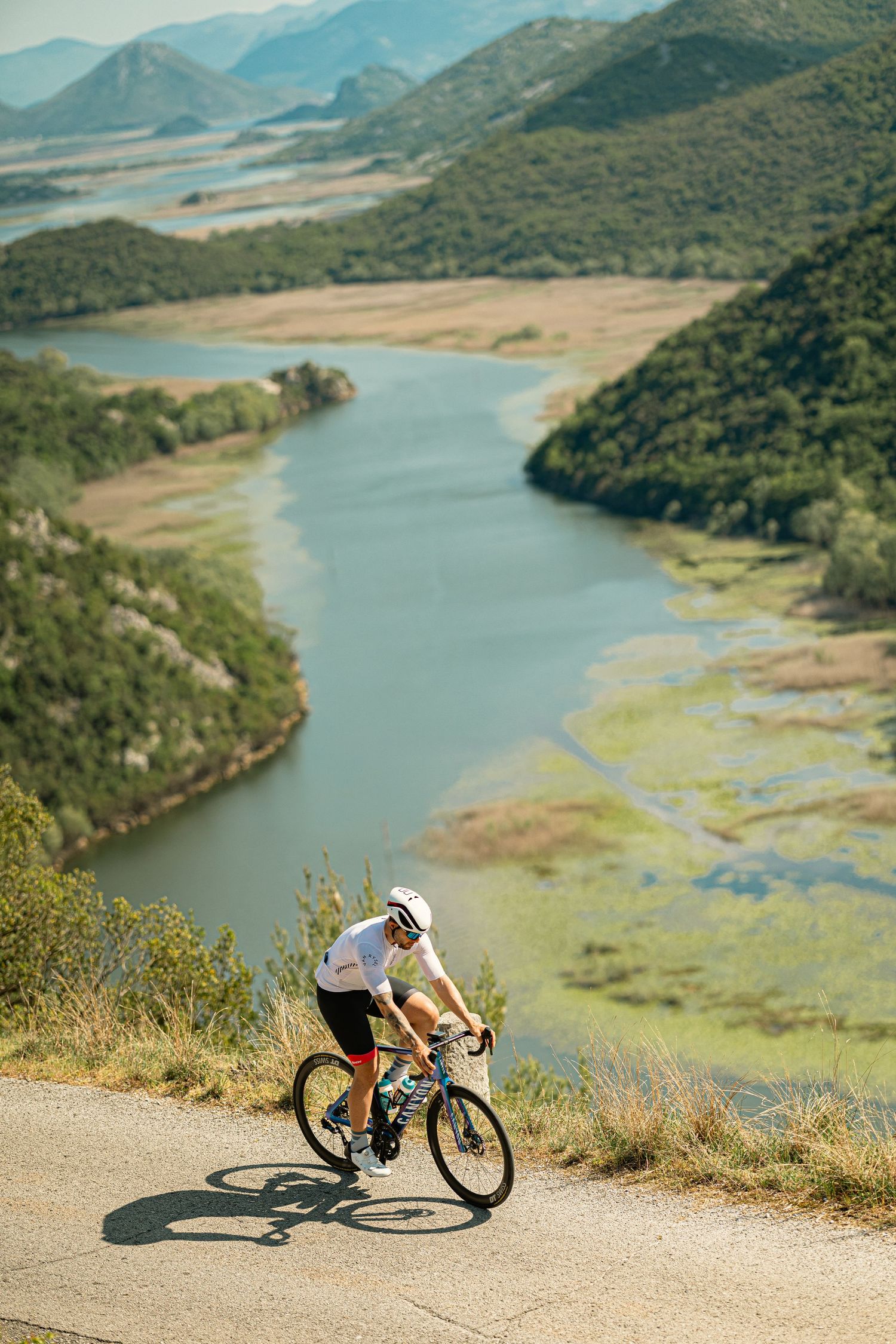 Cyclist in white jersey climbing steep road with scenic river valley and green mountains in background.