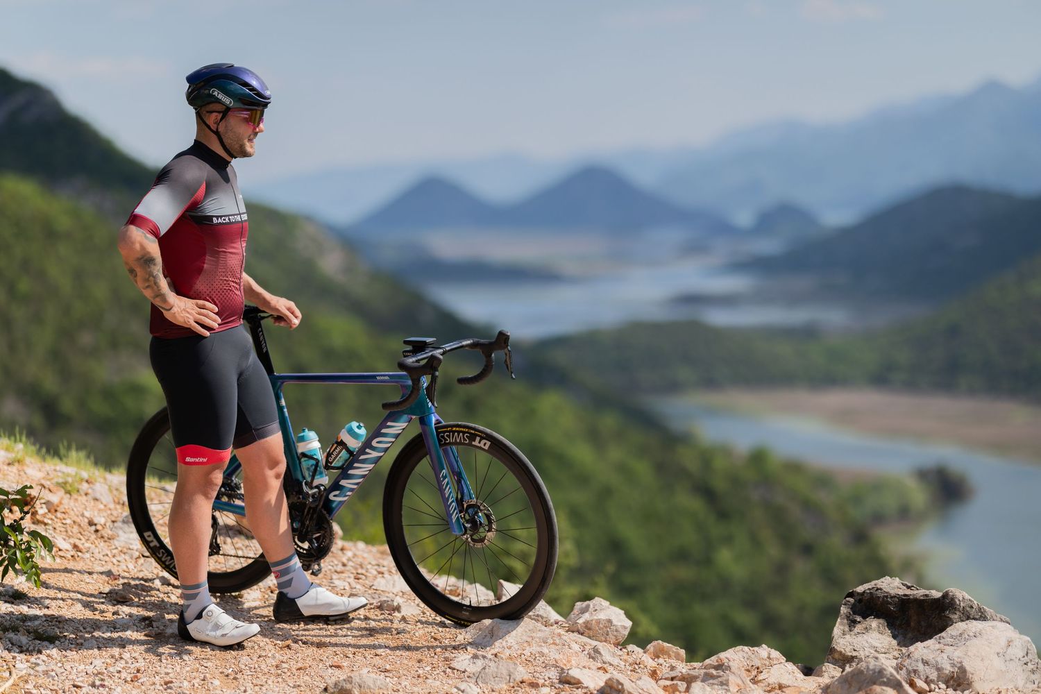 A cyclist in red and white jersey stands beside a blue road bike on a rocky overlook with scenic river and mountain views.