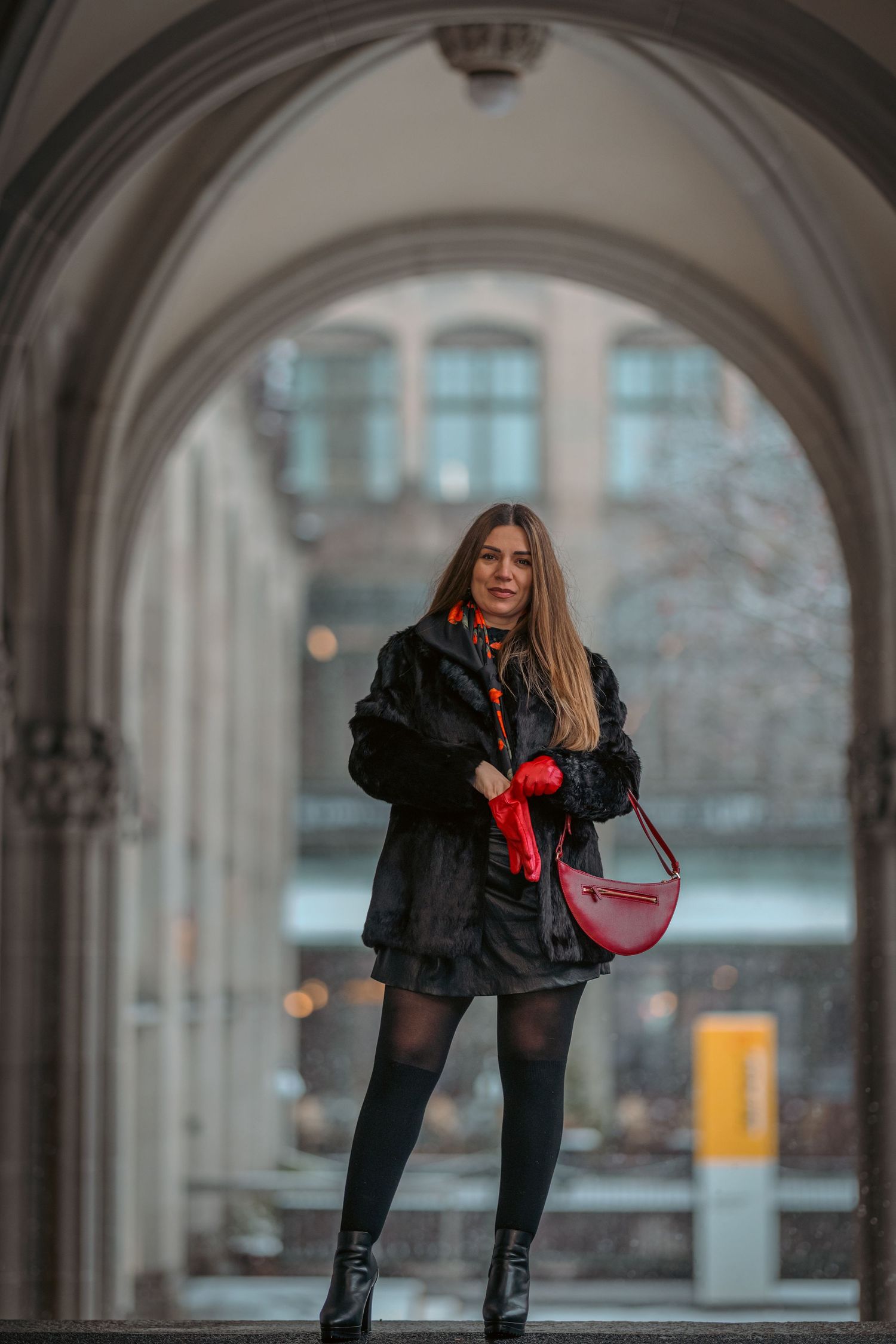 A person in a black jacket and boots poses playfully in front of an arched stone colonnade.