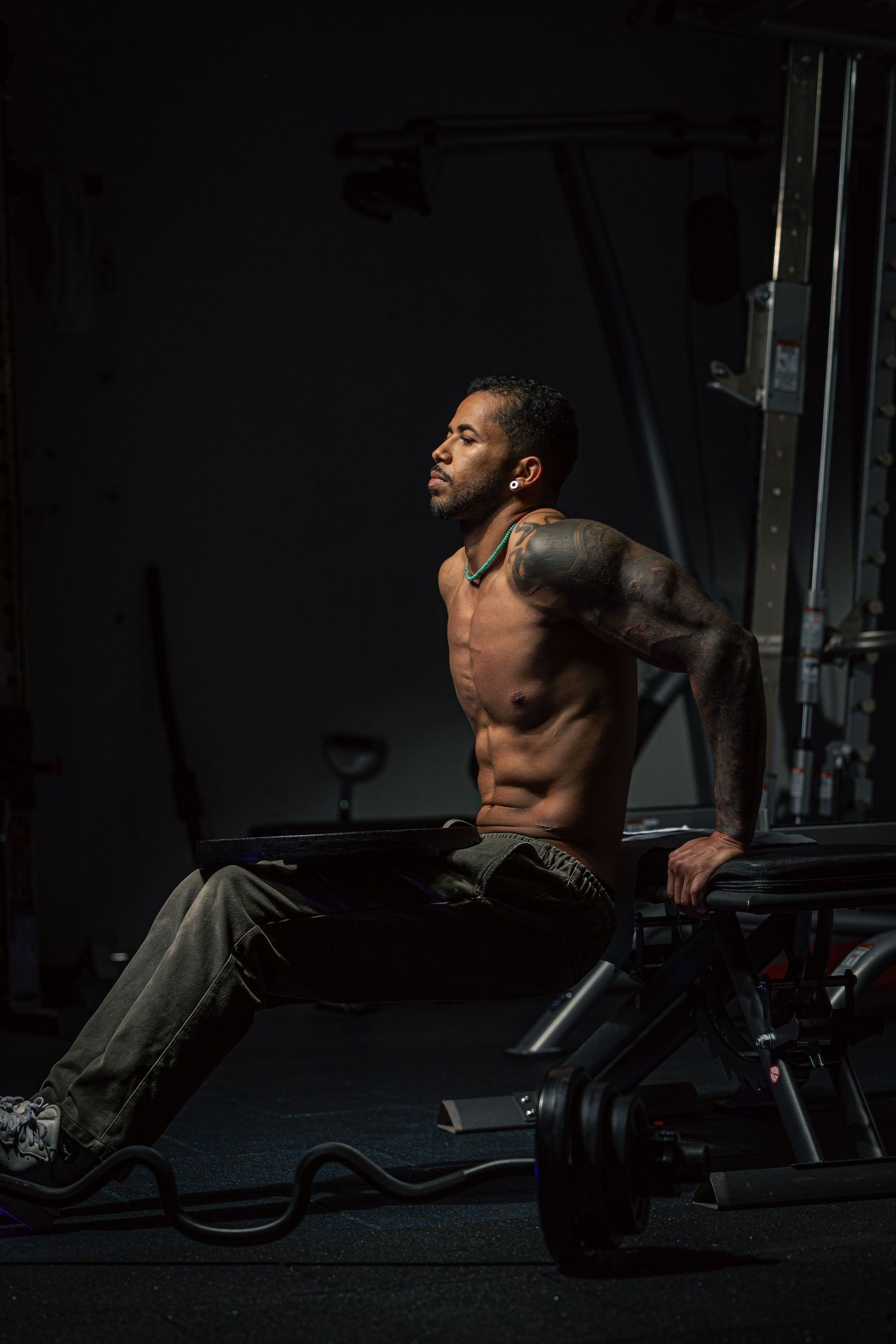 Shirtless tattooed muscular man resting on gym equipment in dramatic dark lighting.