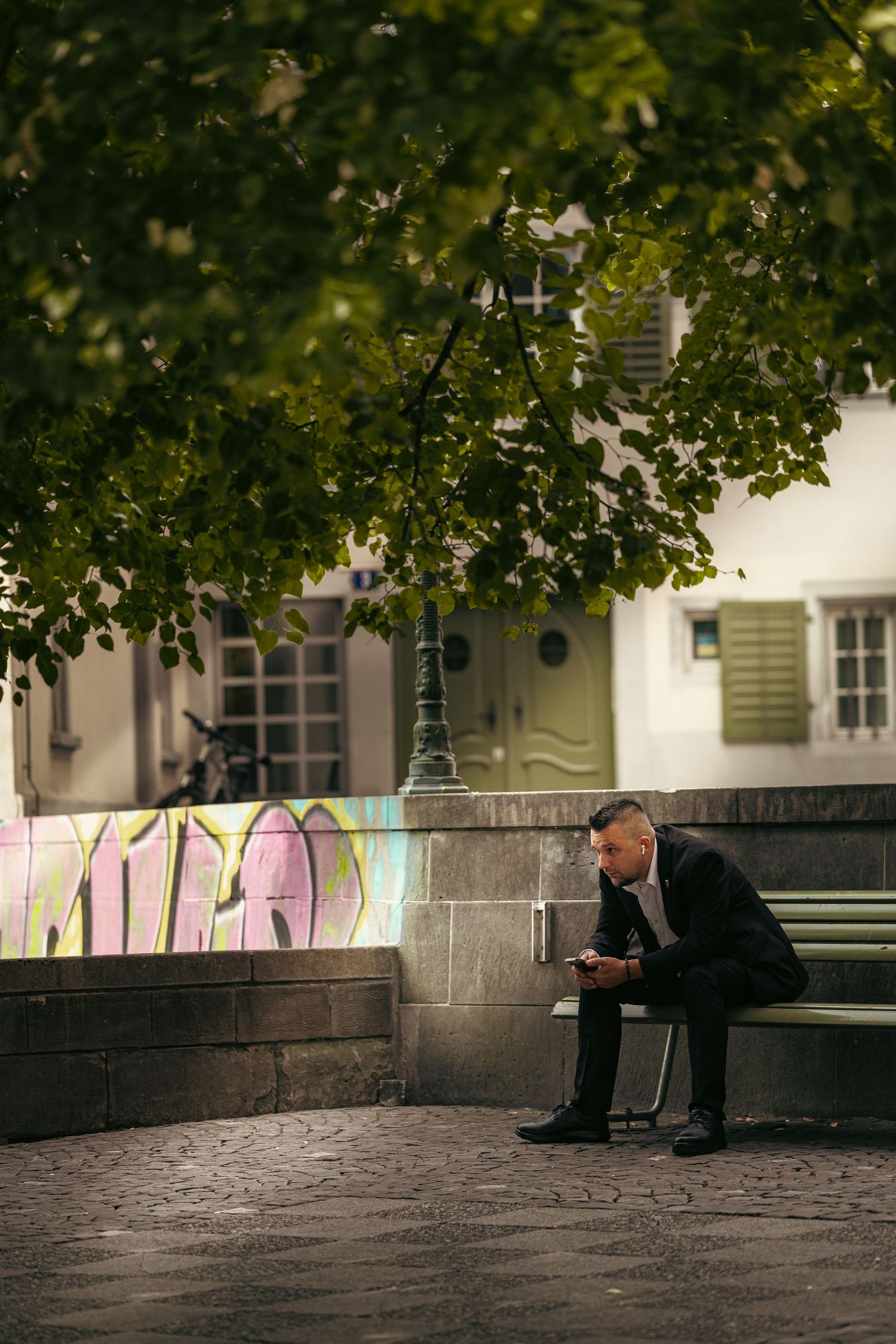 Man in suit sitting on bench using phone outdoors, with graffiti wall and green trees in urban setting.