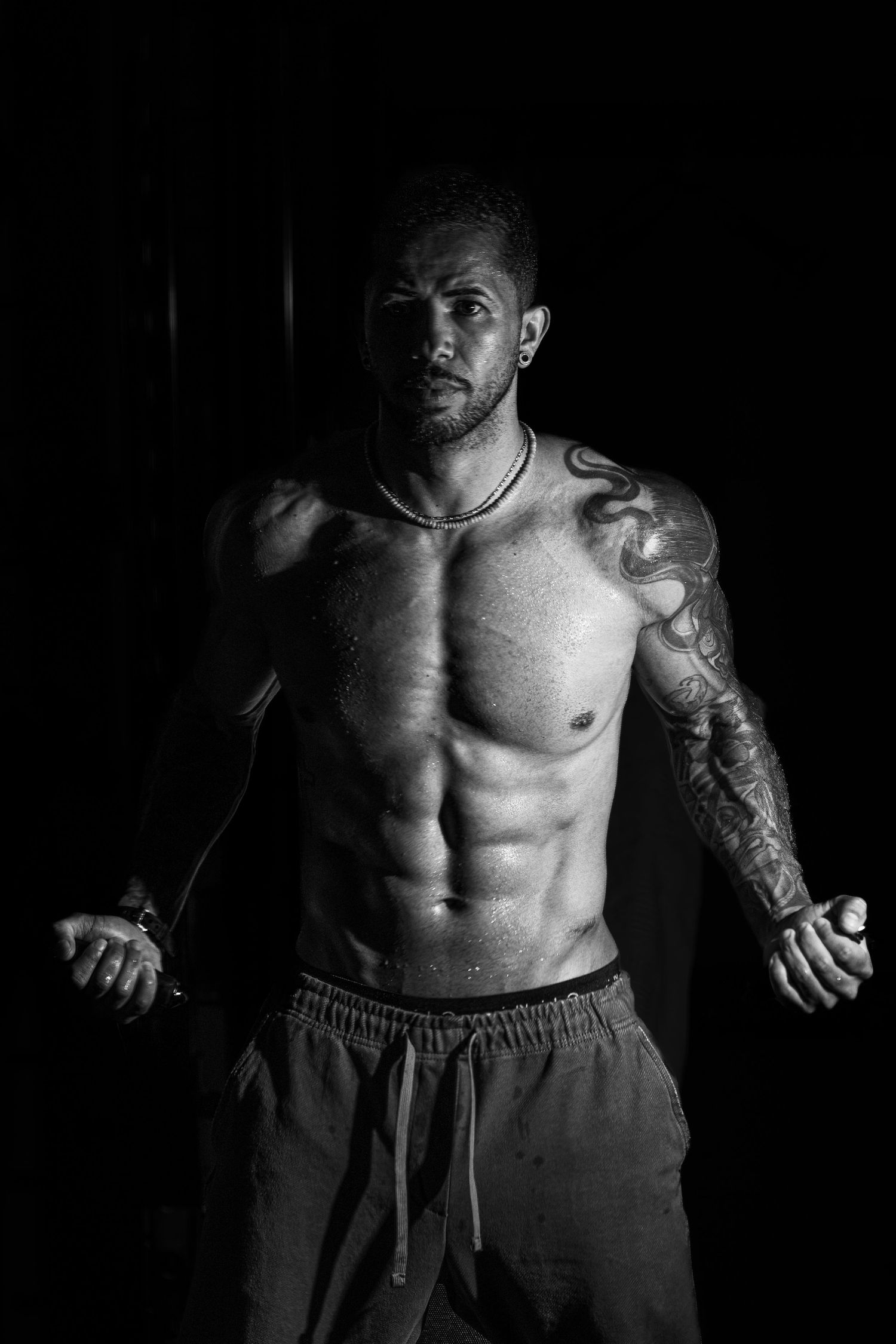 Black and white photo of a muscular shirtless man with tattoos posing against a dark background.